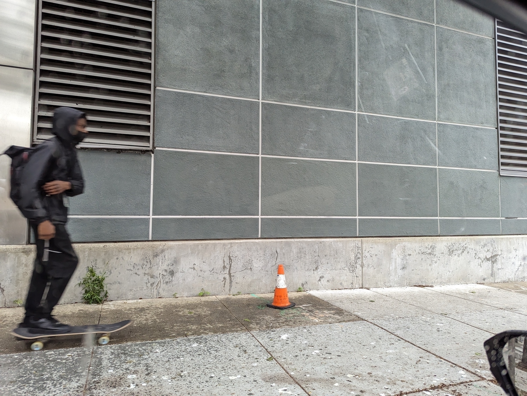 A lean masked hoodie wearing black clad skateboard rider on the leftmost edge of the frame a of about to zip past an orange and white traffic cone Friday, May 5, 2023 while rolling northbound along the western sidewalk in the 1900 block of Franklin Street in downtown Oakland, California.