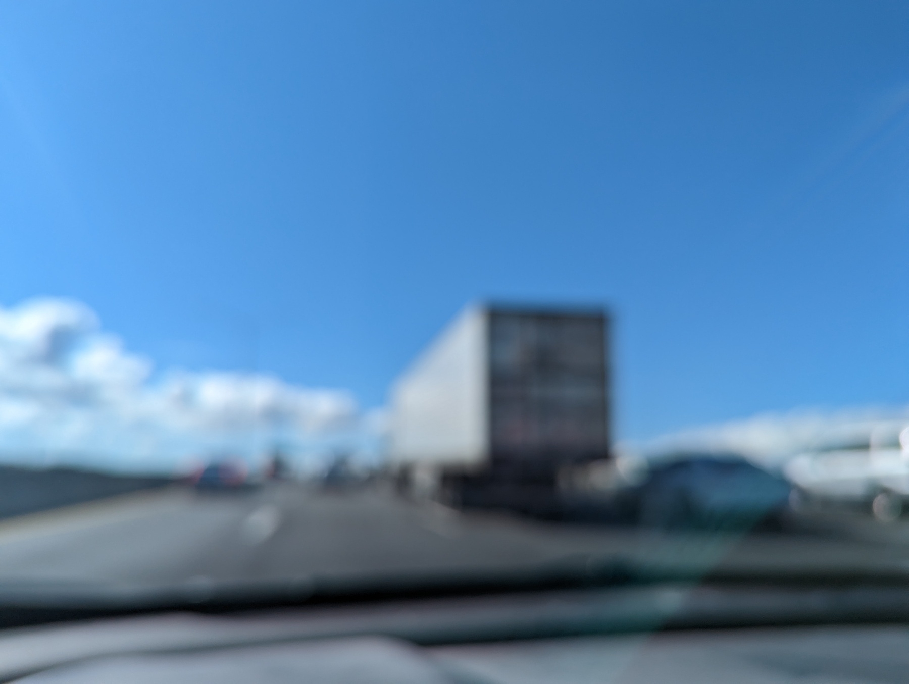 An out of focus image of a white Samia Xpress big rig truck out of Fresno California as seen through a car windshield traveling Thursday March 30th 2023 in westbound Interstate 80 traffic lanes in Albany, California under a bright blue skies in sunny weather with soft puffy white clouds along the horizon
