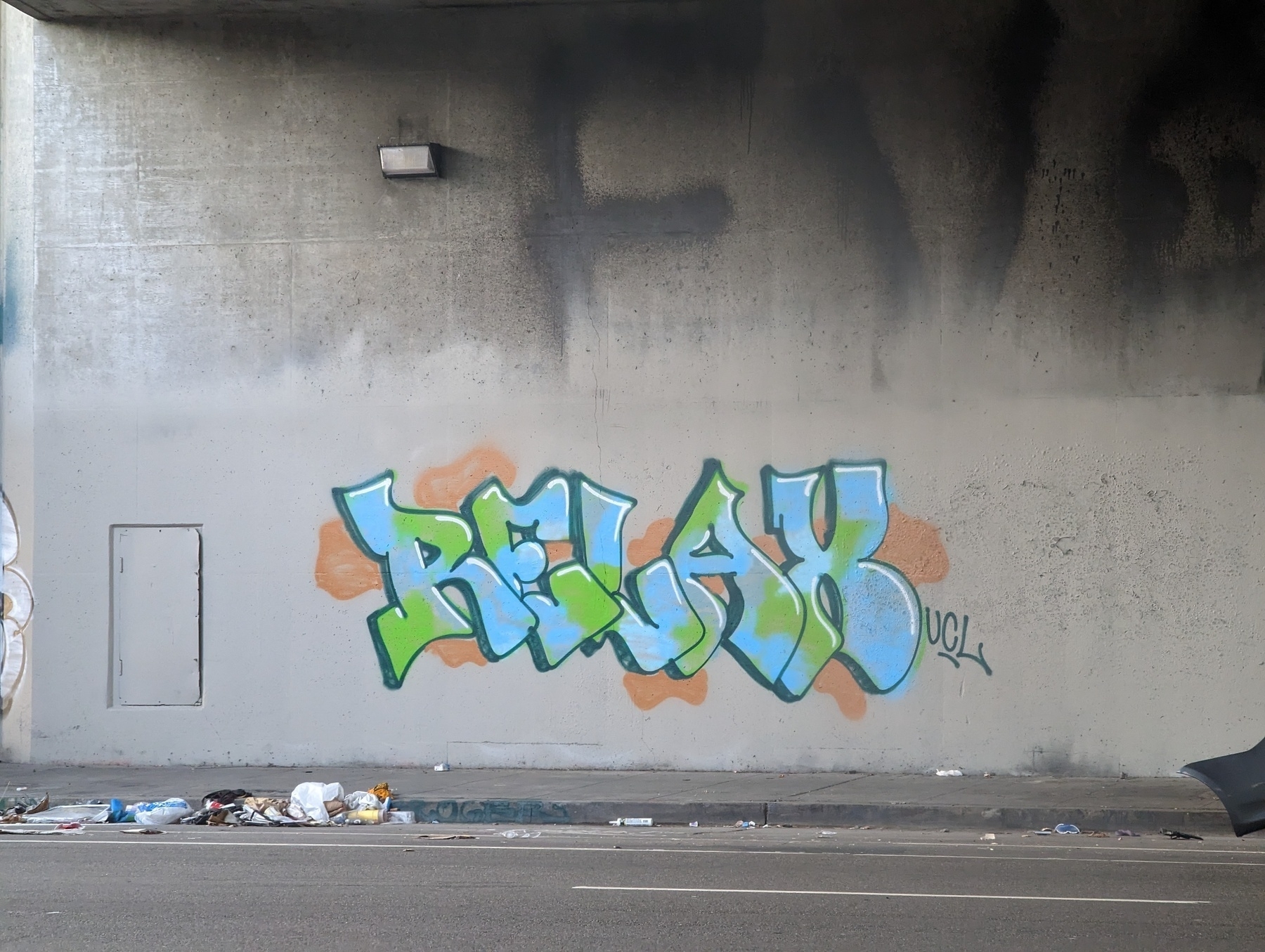 The word "Relax" is sprayed next to its artist tag "UCL" in graffiti with black outlining, blue and green coloring inside and orange patterns around it against a gray concrete wall above a mostly bare sidewalk and curb under an Interstate 980 overpass along 27th Street west of Northgate Avenue Tuesday, May 23, 2023 in Oakland, California.