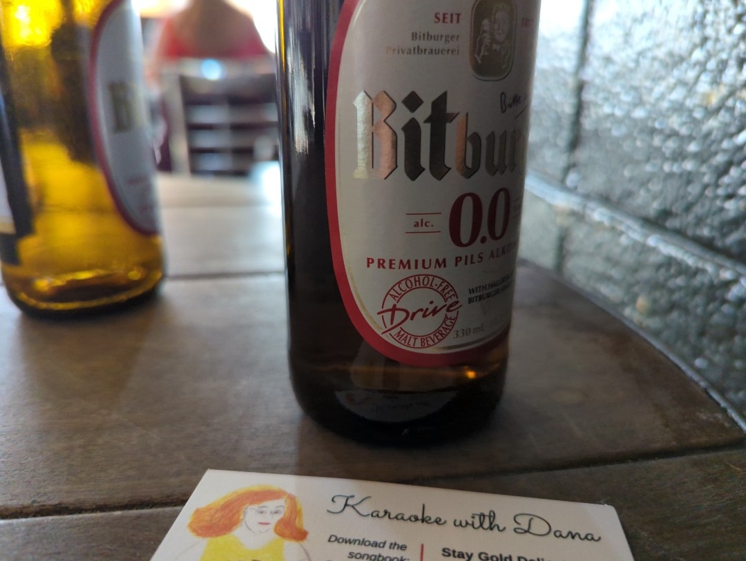 Two brown glass bottles, one close up and another out of focus, of Bitburger premium pilsner nonalcoholic beer sitting on a small round table's wooden top beside a business card advertising karaoke jockey Dana Morrigan's songbook Tuesday, April 11, 2023 in Oakland, California.
