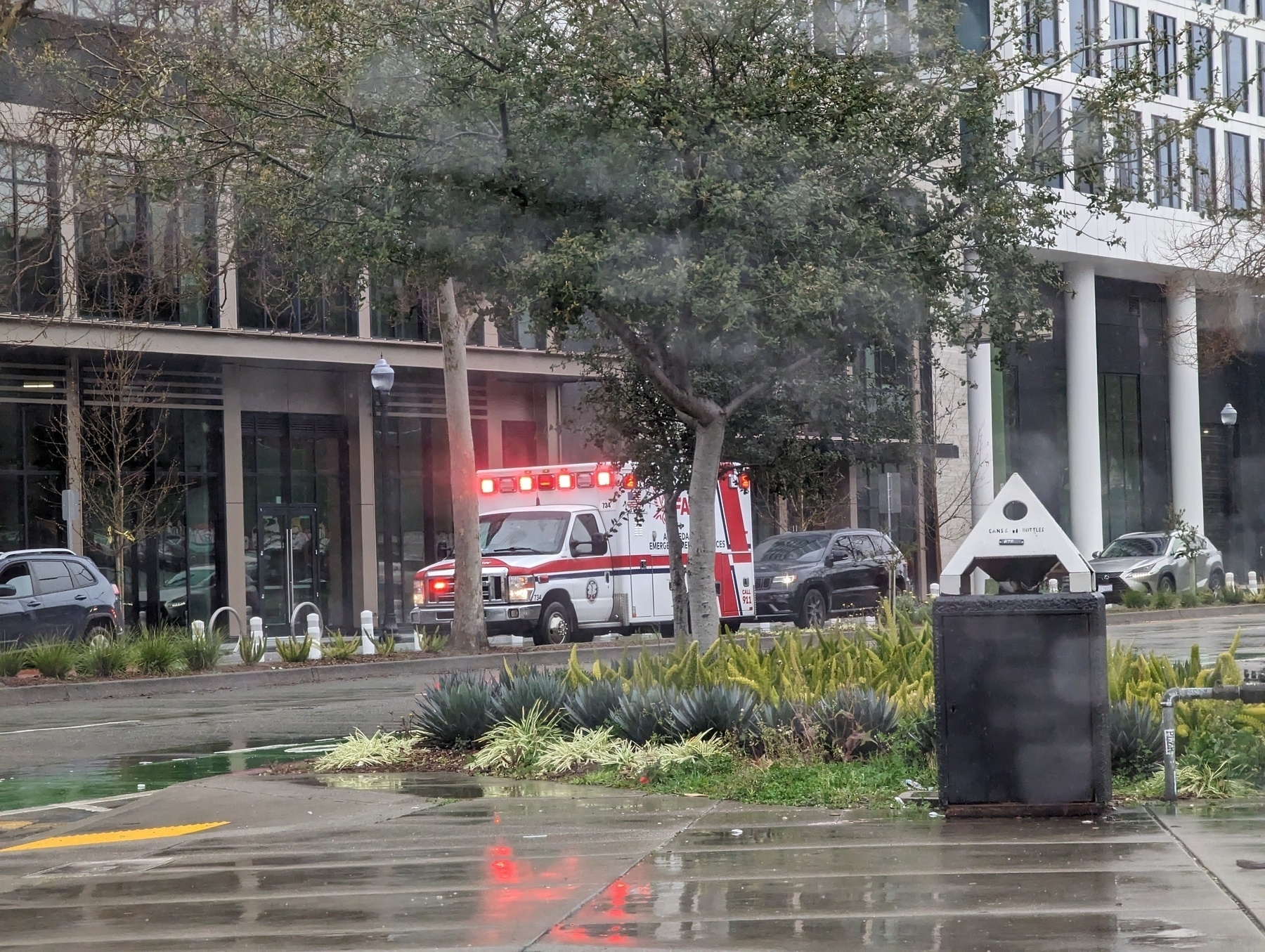 A Falck ambulance with its lights aglow and their glint reflecting on rain wet sidewalks parks along 27th Street just west of Harrison Street around midday Thursday, March 9, 2023 in Oakland, California while waiting to respond to an Oakland Fire Department medical emergency call about a block away in northbound Harrison lanes.
