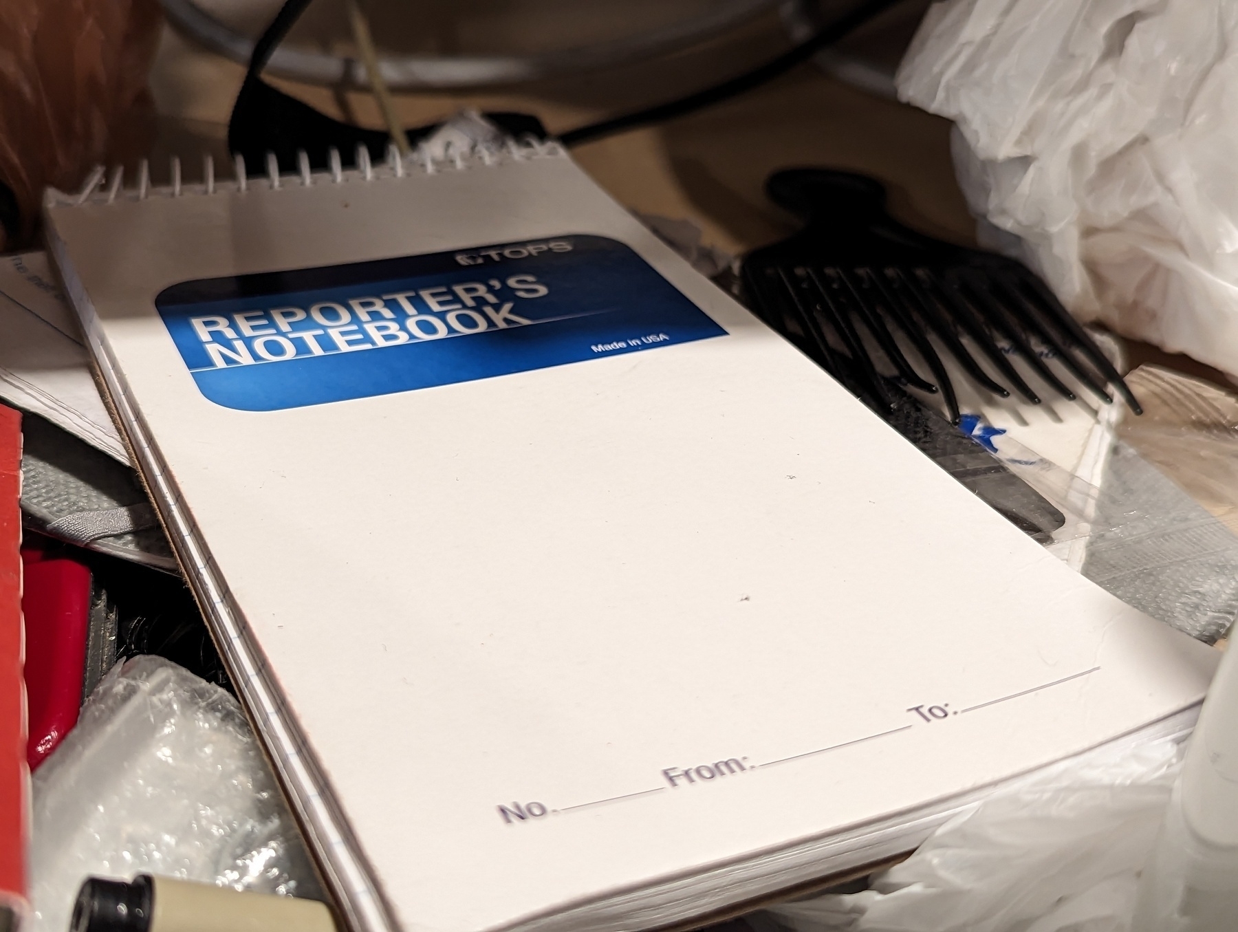A slim white covered spiral bound reporters notebook sits next to a small black pick comb
