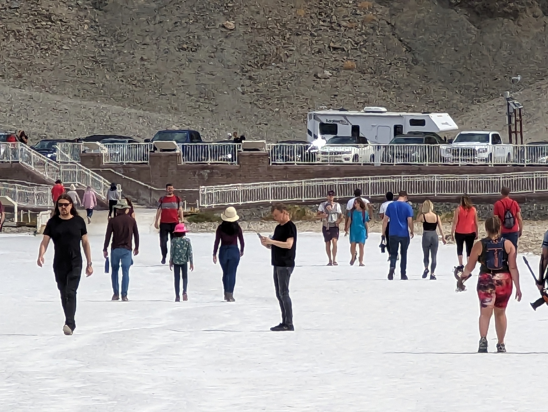 Several dozen people clad in casual clothing walk in warm air and bright sunshine along a salt whitened stretch of open space near a fenced off parking lot full of cars and recreational vehicles Wednesday, March 14, 2023 at Badwater Basin in Death Valley National Park in Death Valley, California. The day's temperatures did not appear to exceed 25 degrees Celsius, or about 76 degrees Fahrenheit.