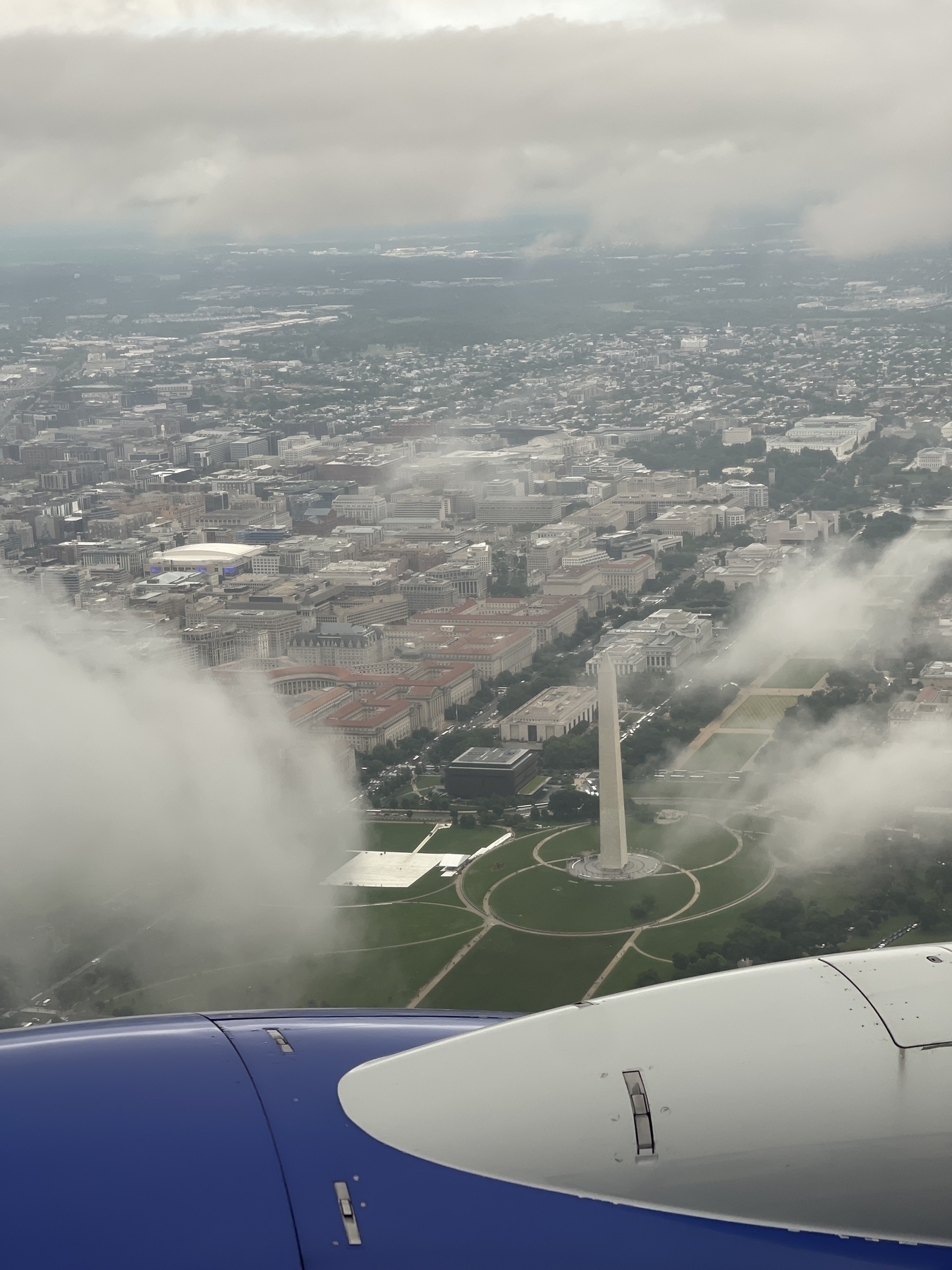 Photograph from an airplane window of the Washington Monument surrounded by clouds.