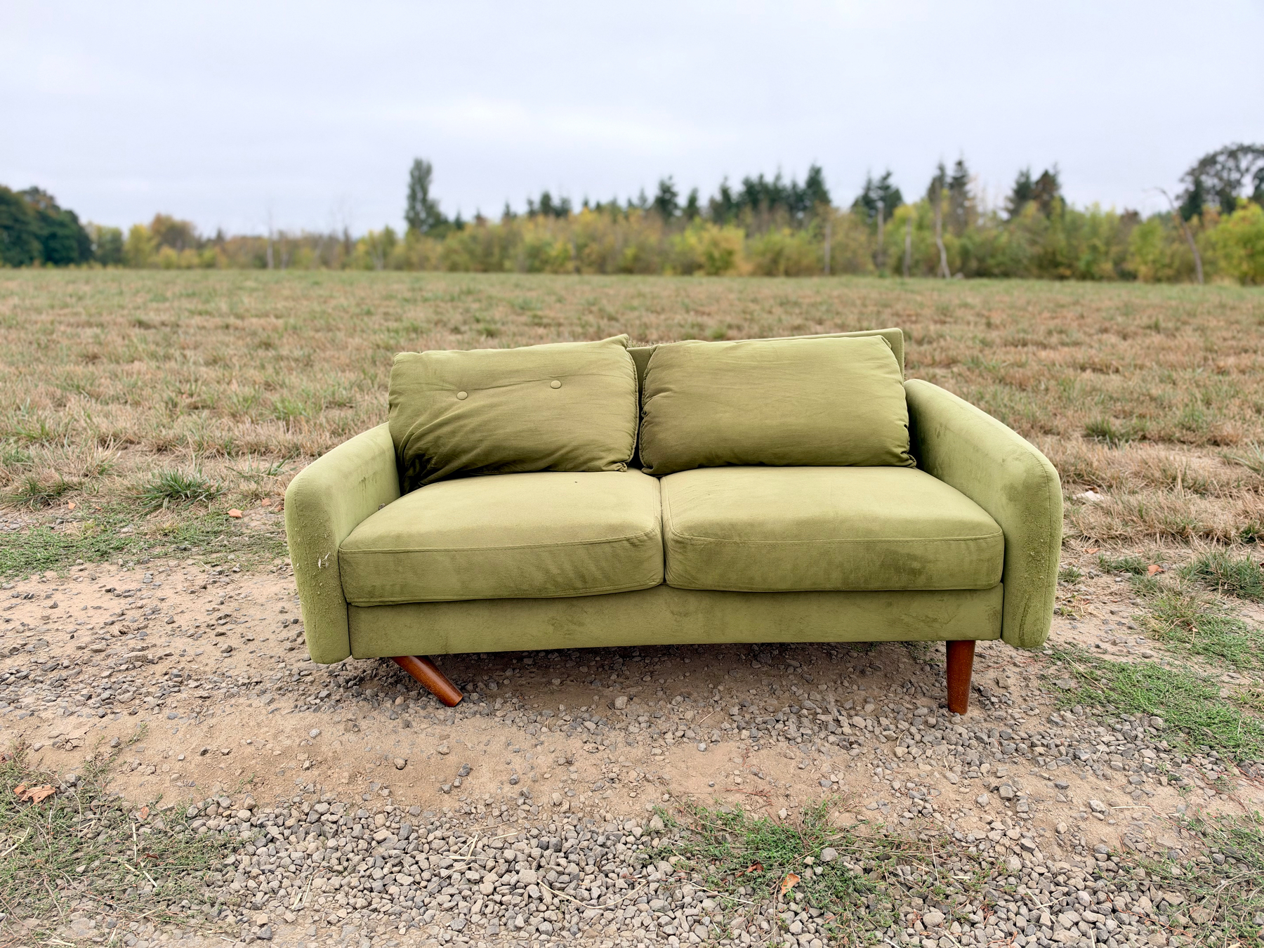 A green sofa is placed on a patch of dirt and gravel in an open field with trees in the background.