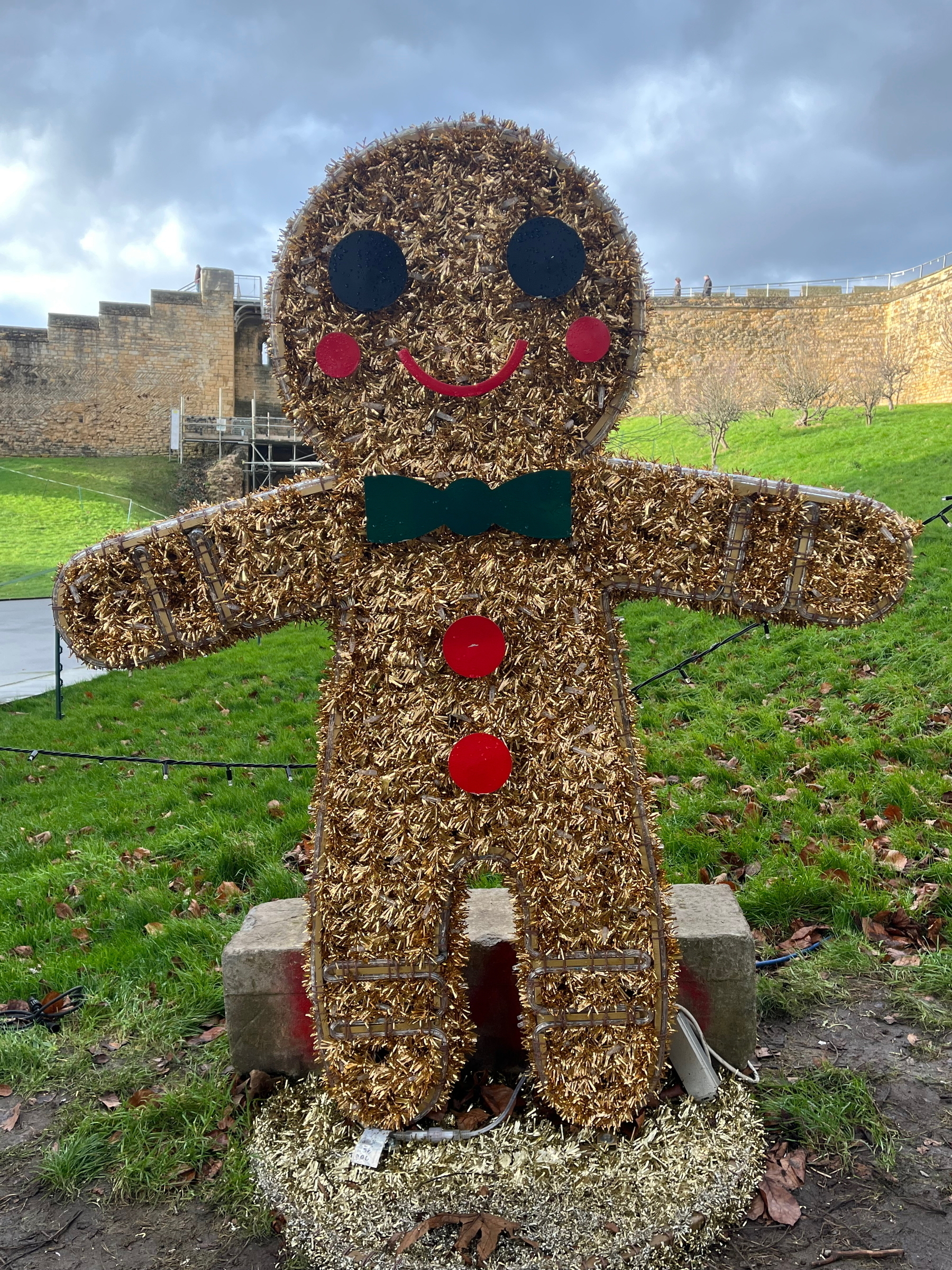 A large, festive gingerbread man with red buttons and a green bow tie stands on grass in front of a stone wall.