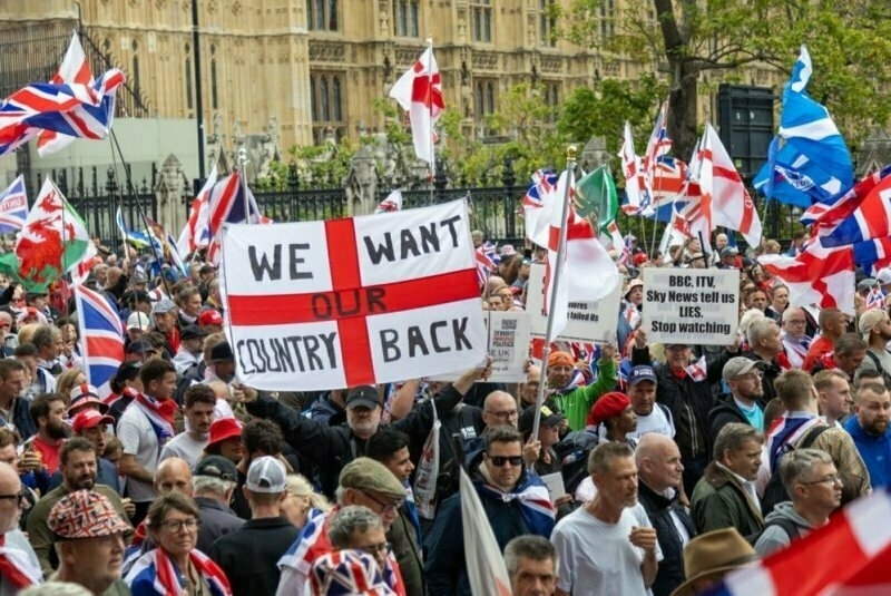 Auto-generated description: A large crowd is gathered, waving flags and holding signs with messages like WE WANT OUR COUNTRY BACK.