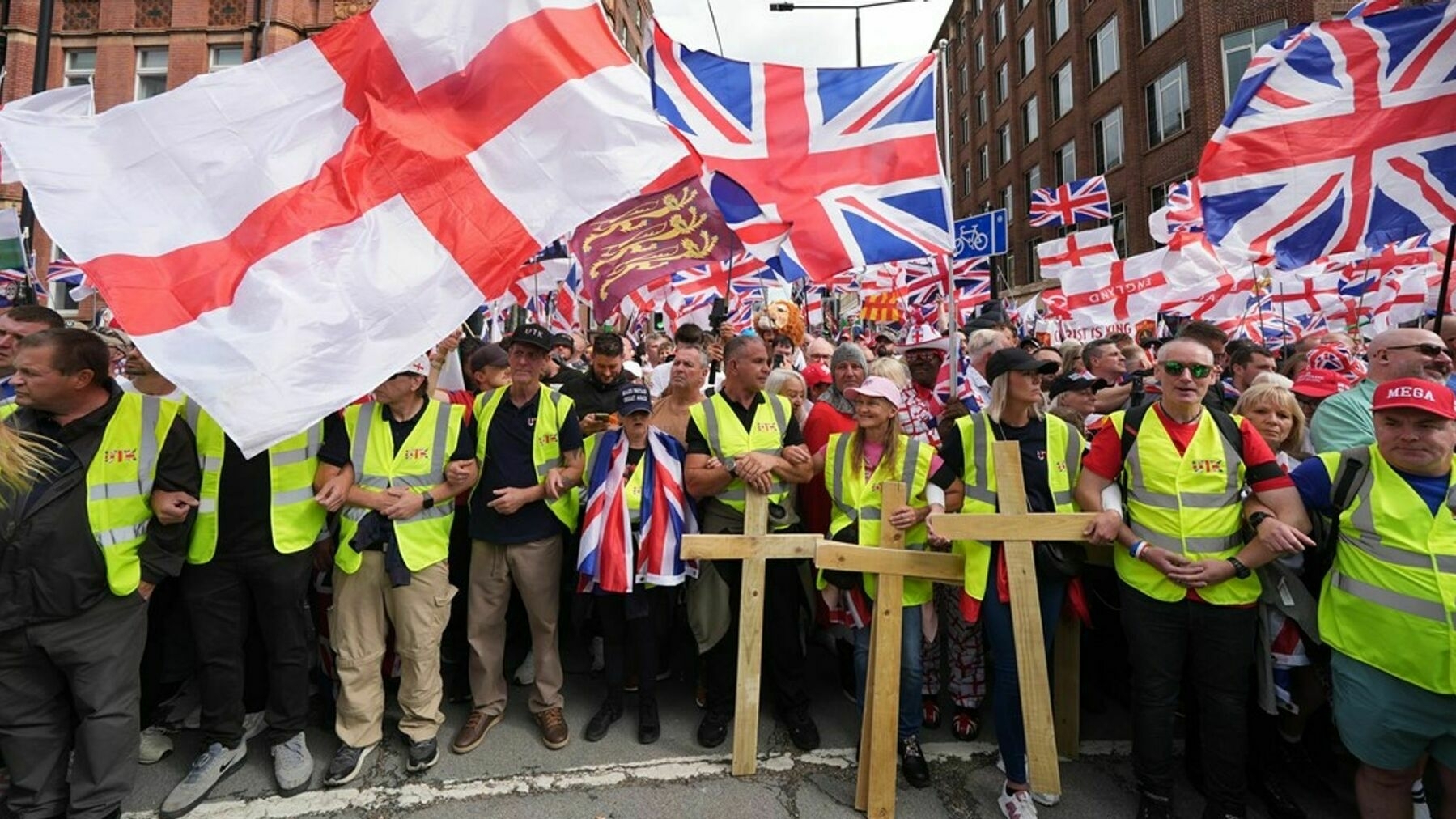 A large group of people, many wearing yellow vests, are holding English and UK flags and wooden crosses during a public gathering or protest.