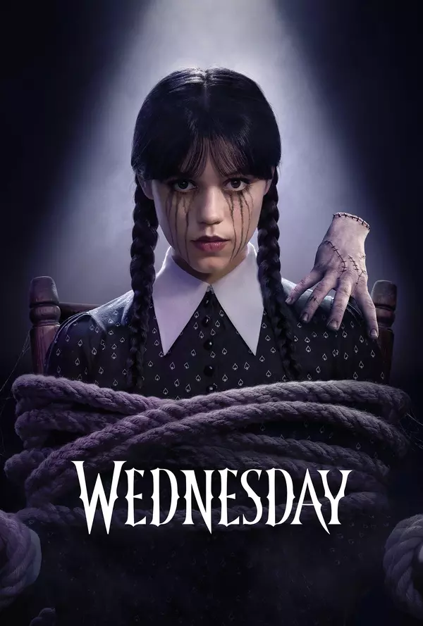 Auto-generated description: A girl with braided hair and a somber expression is seated with a mysterious hand resting on her shoulder, bound by ropes, under a dramatic spotlight with the title Wednesday below.