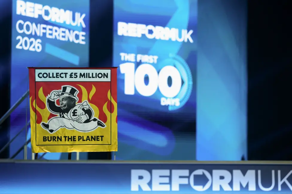 Auto-generated description: A banner displaying Collect £5 million, Burn the Planet is positioned in front of a stage backdrop for the Reform UK Conference 2026, emphasizing The First 100 Days.