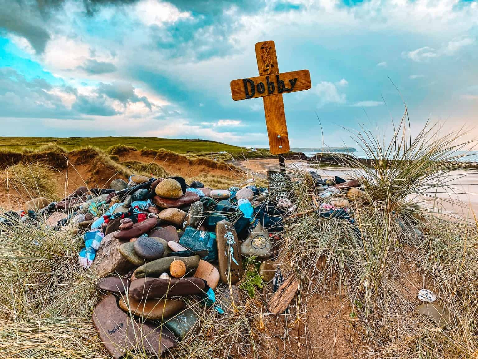 A wooden cross with the name Dobby marks a memorial site adorned with various stones, small objects, and trinkets on a sandy, grassy beach.