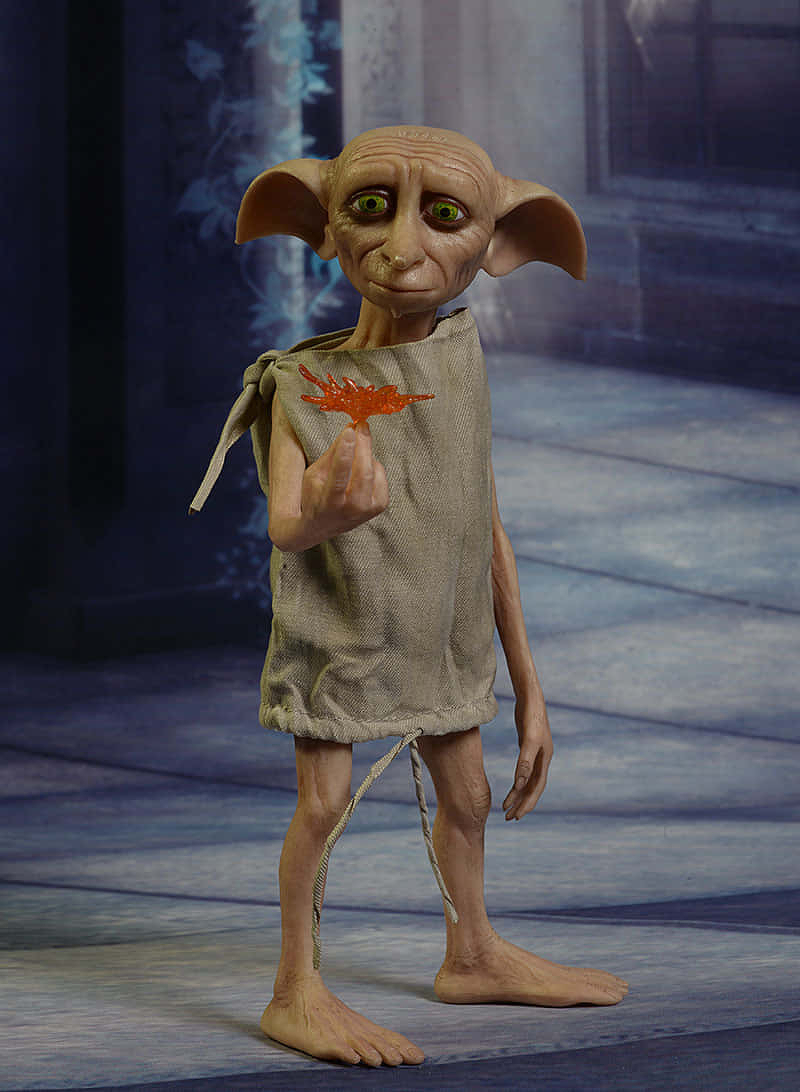 Dobby, a small creature with large ears and green eyes is holding an orange leaf while wearing a simple beige tunic.