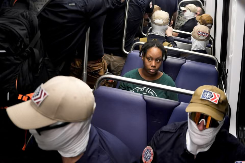 A young Black woman sits alone on a subway train surrounded by Patriot Front members wearing face coverings and uniforms.