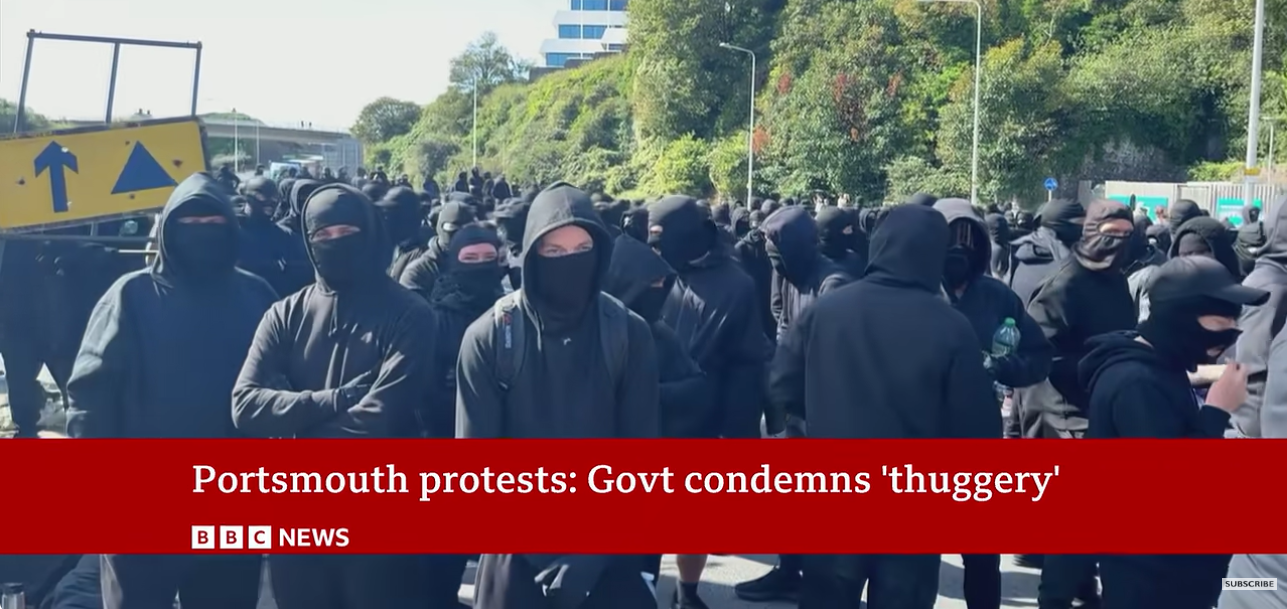 Auto-generated description: A large group of protesters dressed in black and wearing face coverings are gathered on a street, with a BBC News headline stating Portsmouth protests: Govt condemns 'thuggery'.
