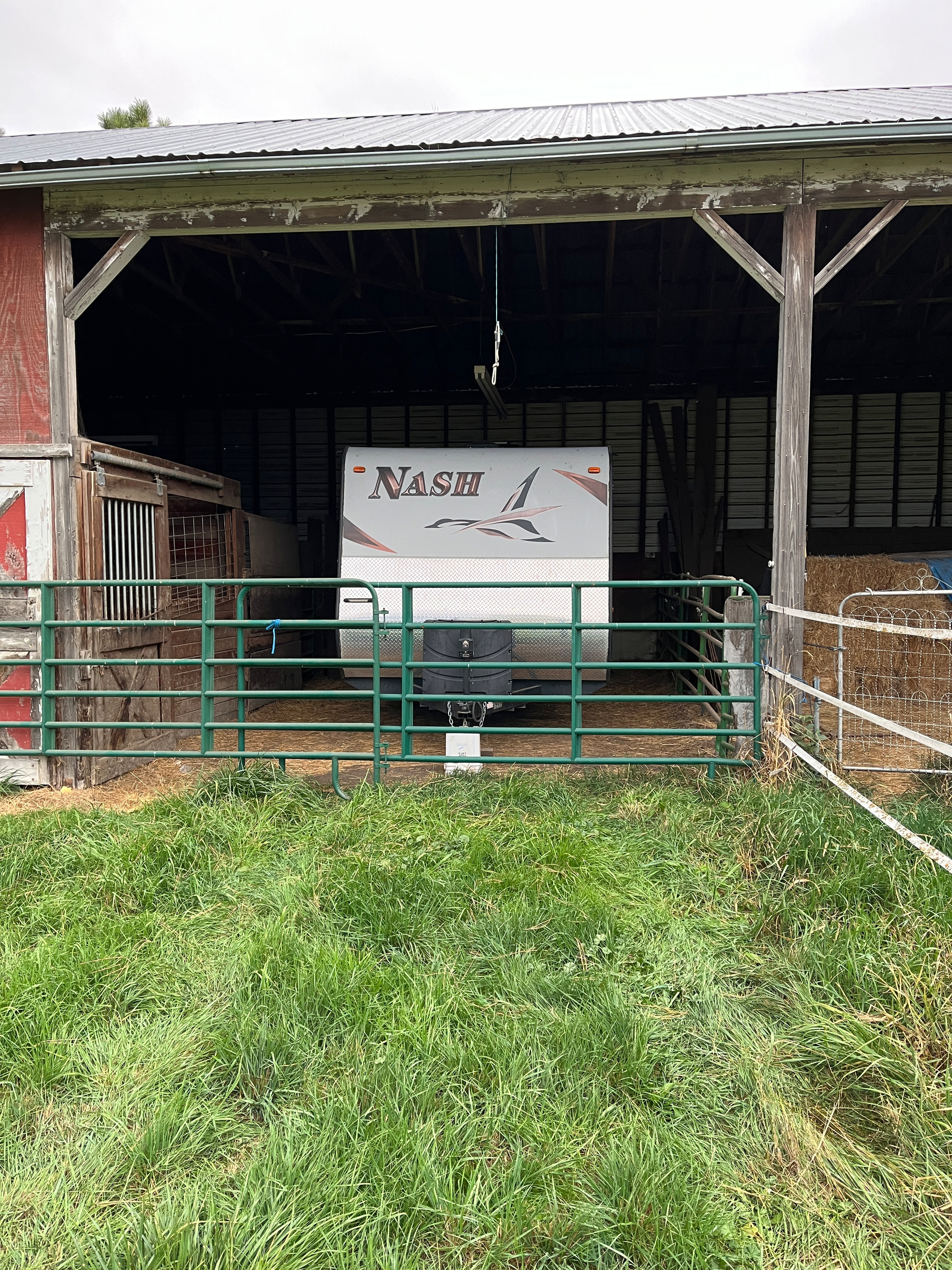 A Nash RV trailer is stored inside a barn behind a green metal gate with grass in the foreground.