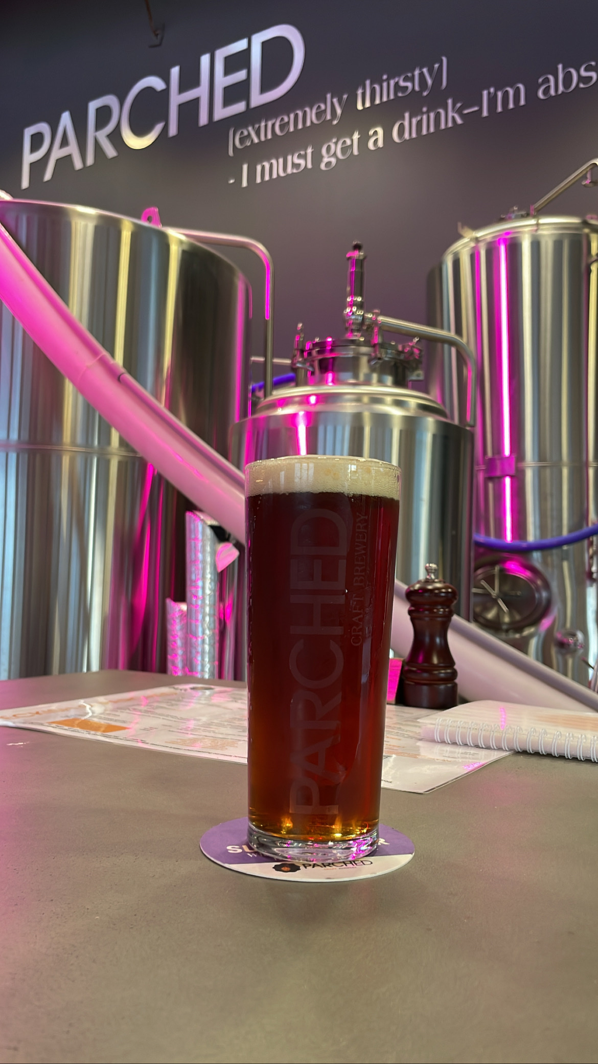 A schooner of beer in front of stainless steel brewing vats. The wall in the background says PARCHED (extremely thirsty).