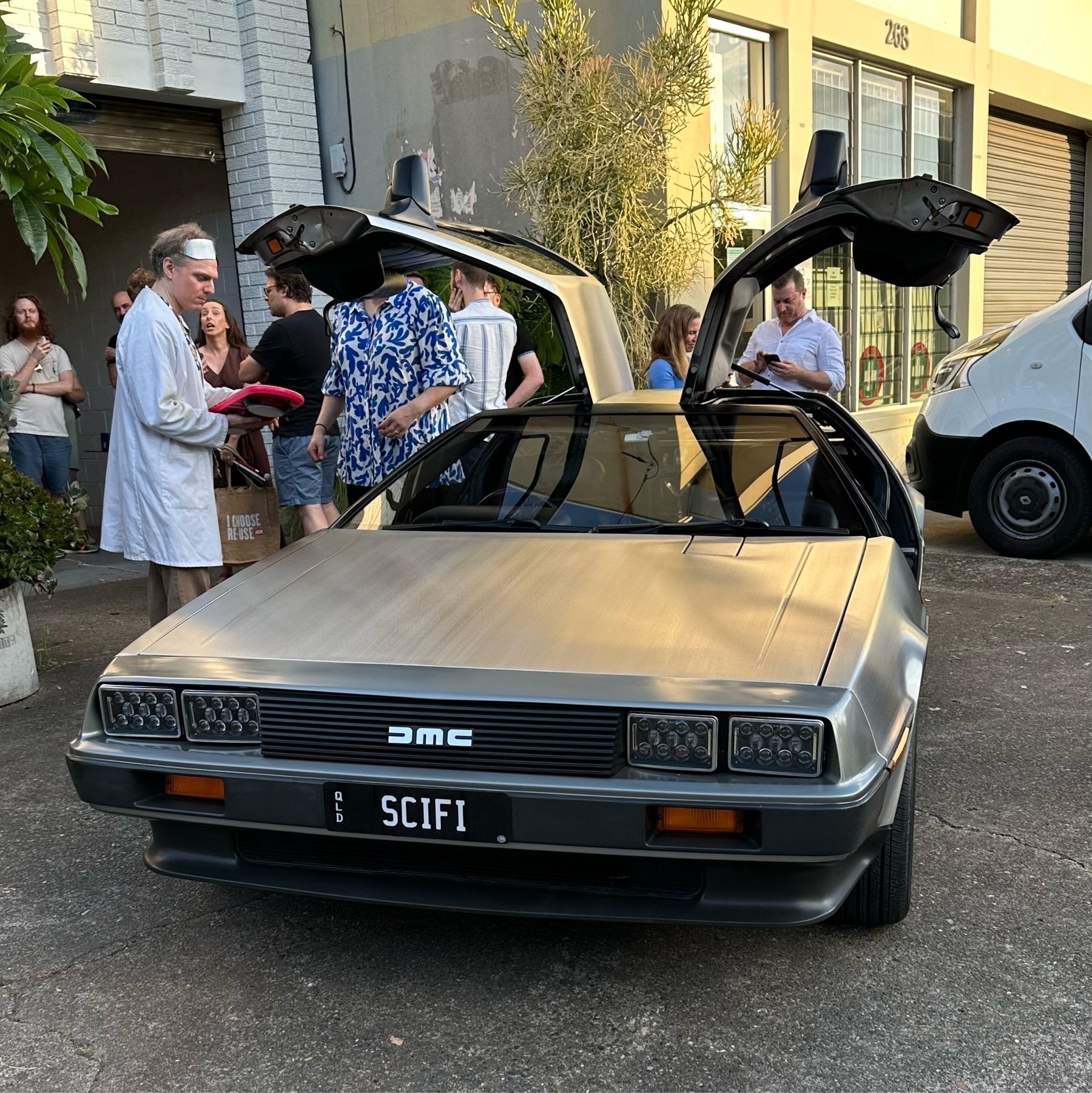 A DeLorean car parked with the winged doors raised up. The licence plate is SCIFI and a man dressed as the Doc from Back to the Future stands nearby.