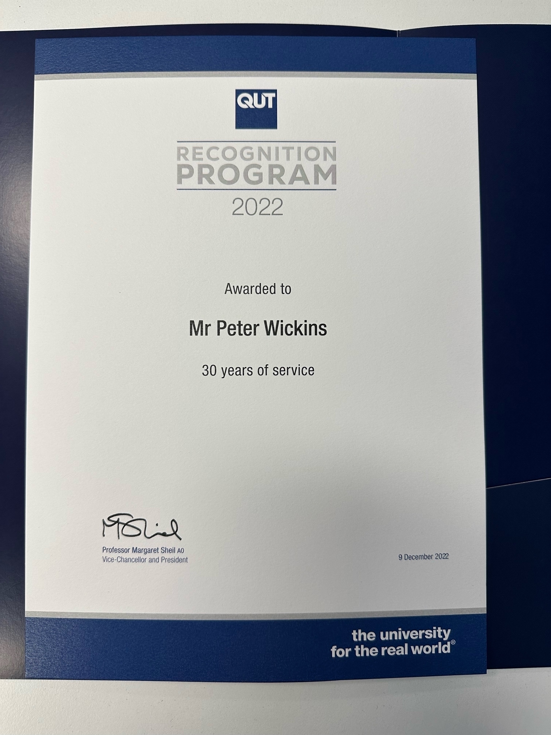 A white certificate bordered in blue. At the top is the word QUT inside a blue box with “Recognition Program 2022” underneath. In the middle is “Awarded to Mr Peter (surname is blocked out) for 30 years of service”. Bottom left has the name and signature of the Vice-Chancellor. Bottom right is 9 December 2022.