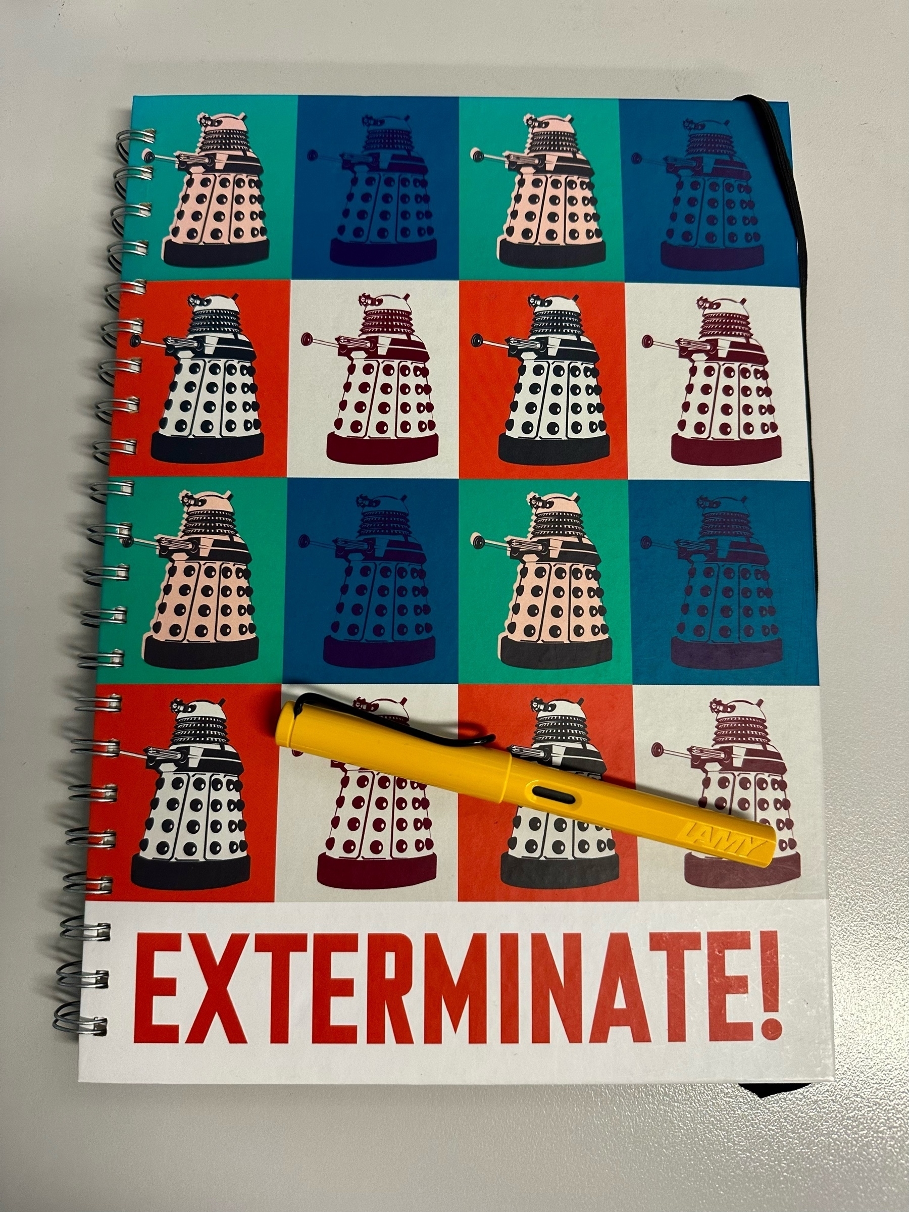 A spiral bound A4 hardcover notebook. There is a 4x4 grid of squares of different colours with a Dalek inside each one. Across the bottom is Exterminate! in red capital letters. A yellow pen is sitting on the book.