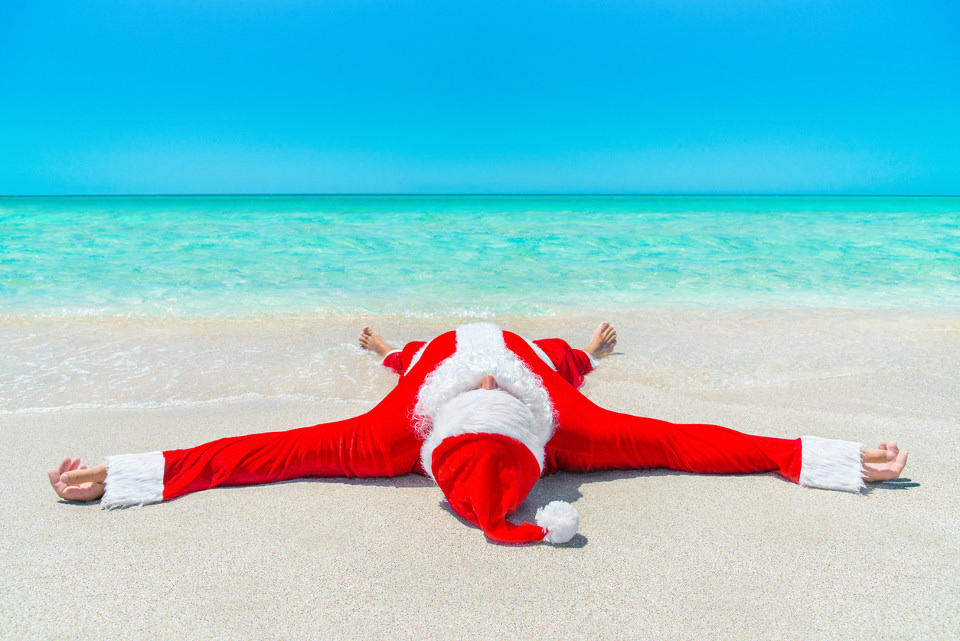 Santa spreadeagled on his back on a beach. His pants a rolled up and his feet are just in the water. The sea is a clear blue and beach a bright white.