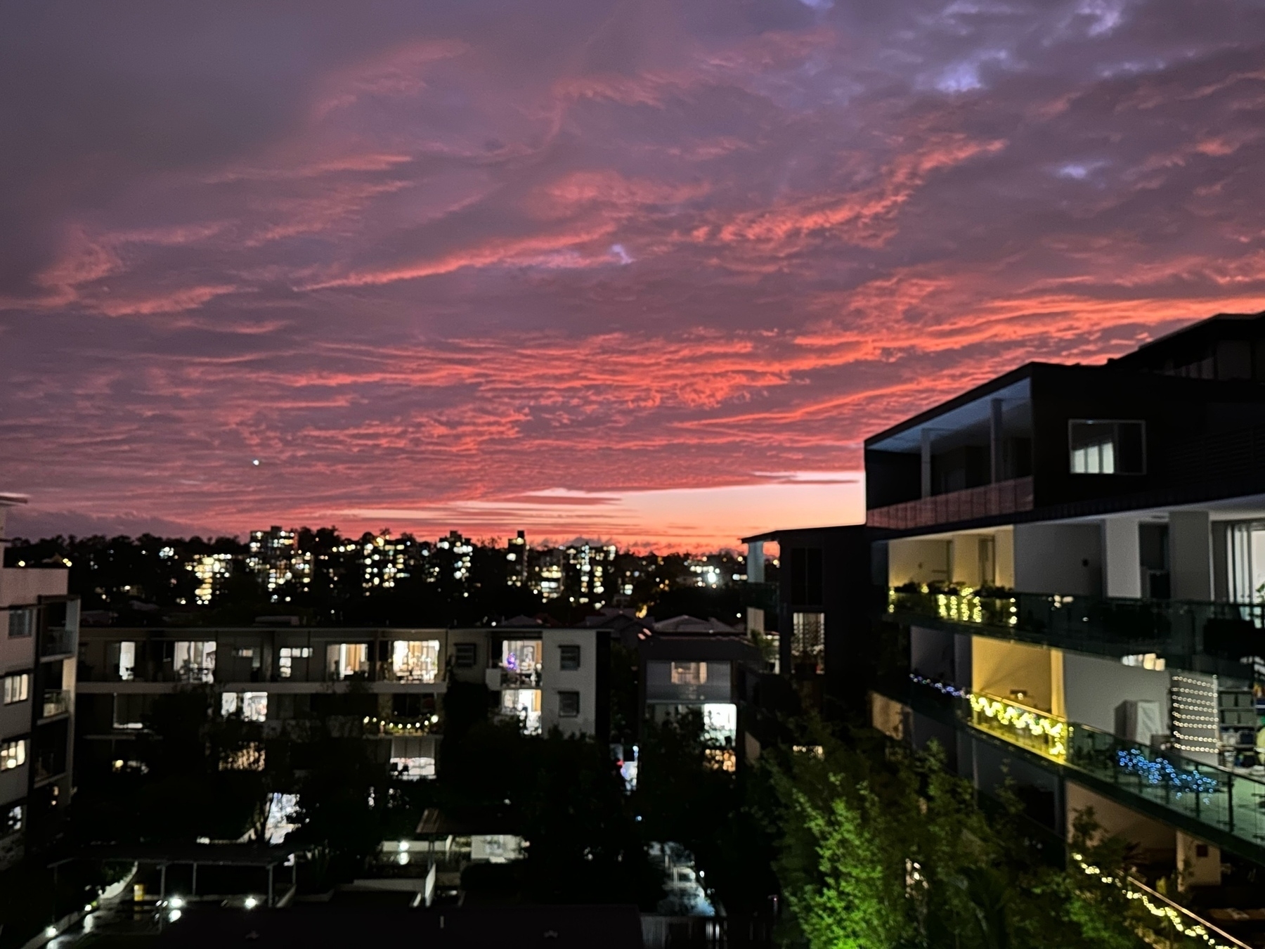 A view from an apartment balcony over some low buildings, another apartment building to the right. The sky is full of clouds that are a deep dark red, almost purple, colour.