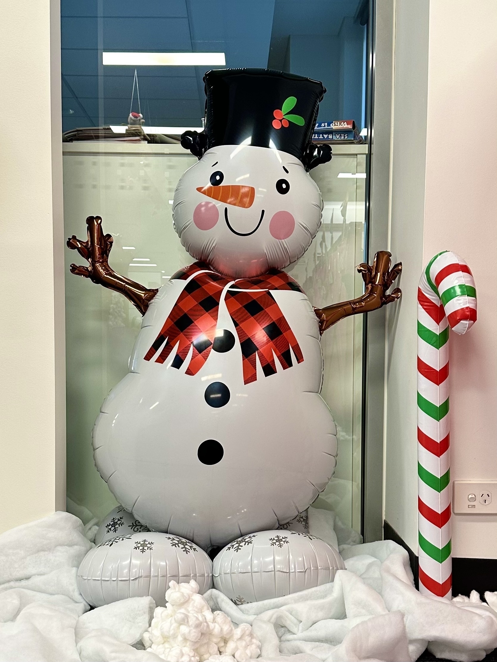 An inflatable snowman about 1 metre high standing against a glass panel. The snowman has a black top hat, red scarf, carrot for a nose and branches for arms. An inflatable red, white and green candy cane is standing nearby.