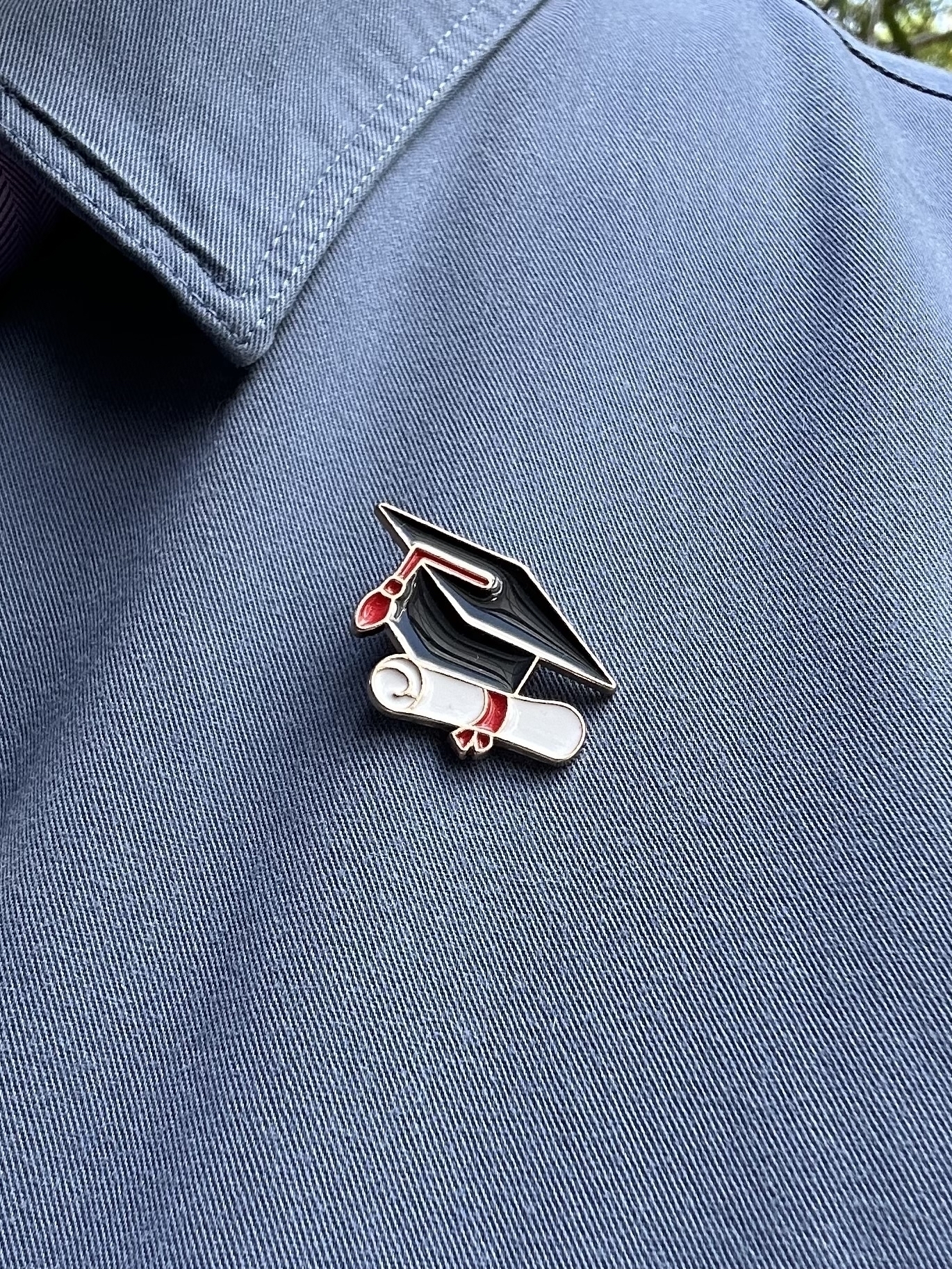 A lapel badge pinned on a blue shirt. The badge is a black mortar board with red tassle above a rolled up white document with a red ribbon around it.