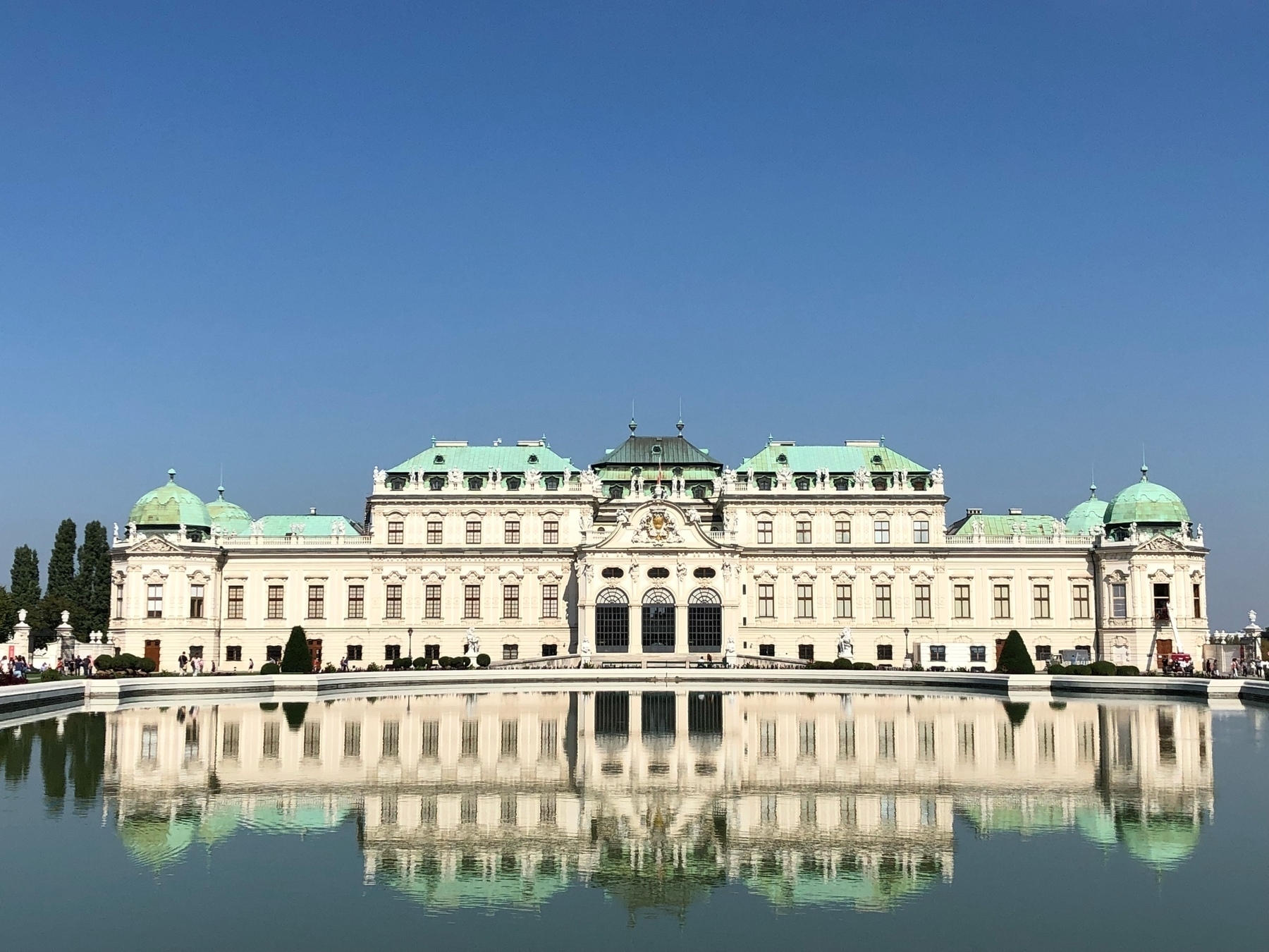 A large white baroque palace with green roof, two main levels and a smaller third and fourth level in the middle third. The palace is clearly reflected in the very large pond in front.