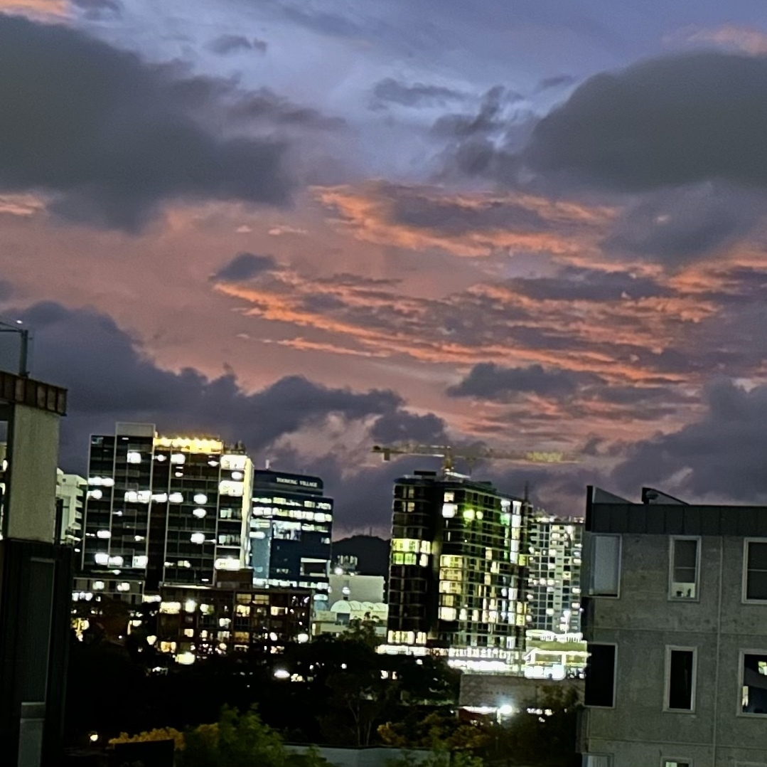 Looking towards a city scape. Apartment buildings shining brightly with their windows lit up and above them a red sky and red/grey tinged clouds.