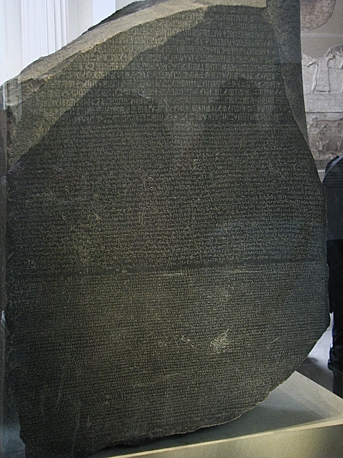 A large black stone with a smooth front surface but rough edges where it’s been broken. There are three different languages carved on the stone. It’s inside a glass case and the reflections of people viewing it are visible in the glass.