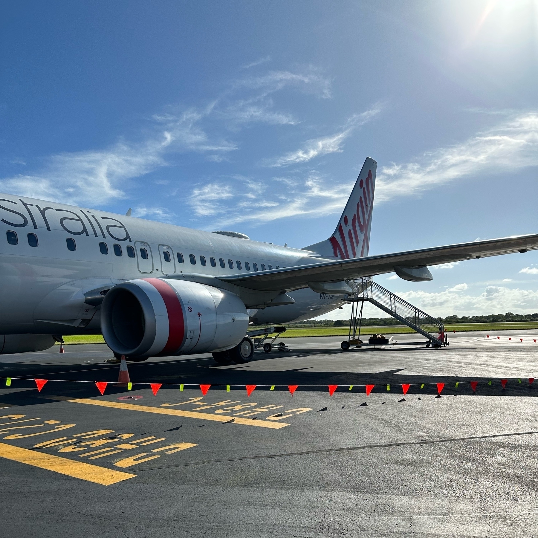 A Virgin Australia plane on the tarmac, one engine and wing along with the tail is visible. A boarding stairs are at the rear door. A bright blue sky in the background.