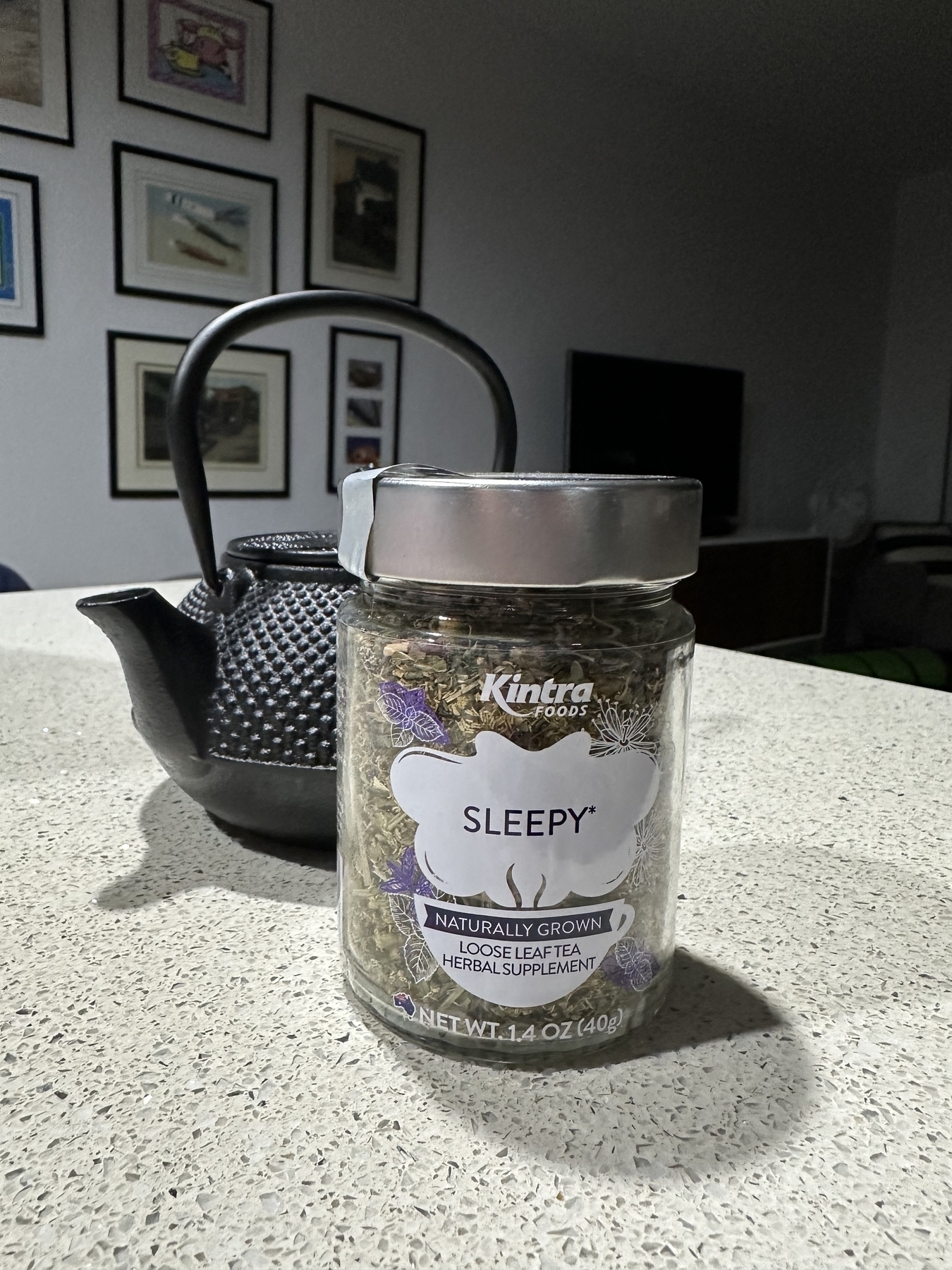 A glass jar of herbal tea sitting in front of a black teapot on a kitchen bench. The tea is “Sleepy” by Kintra Foods.