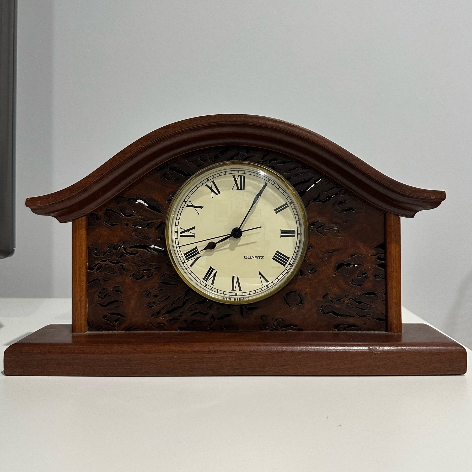 A round clock face showing 8:05 set into carved wood. The shape is a classic mantel clock with flat base and curved top.