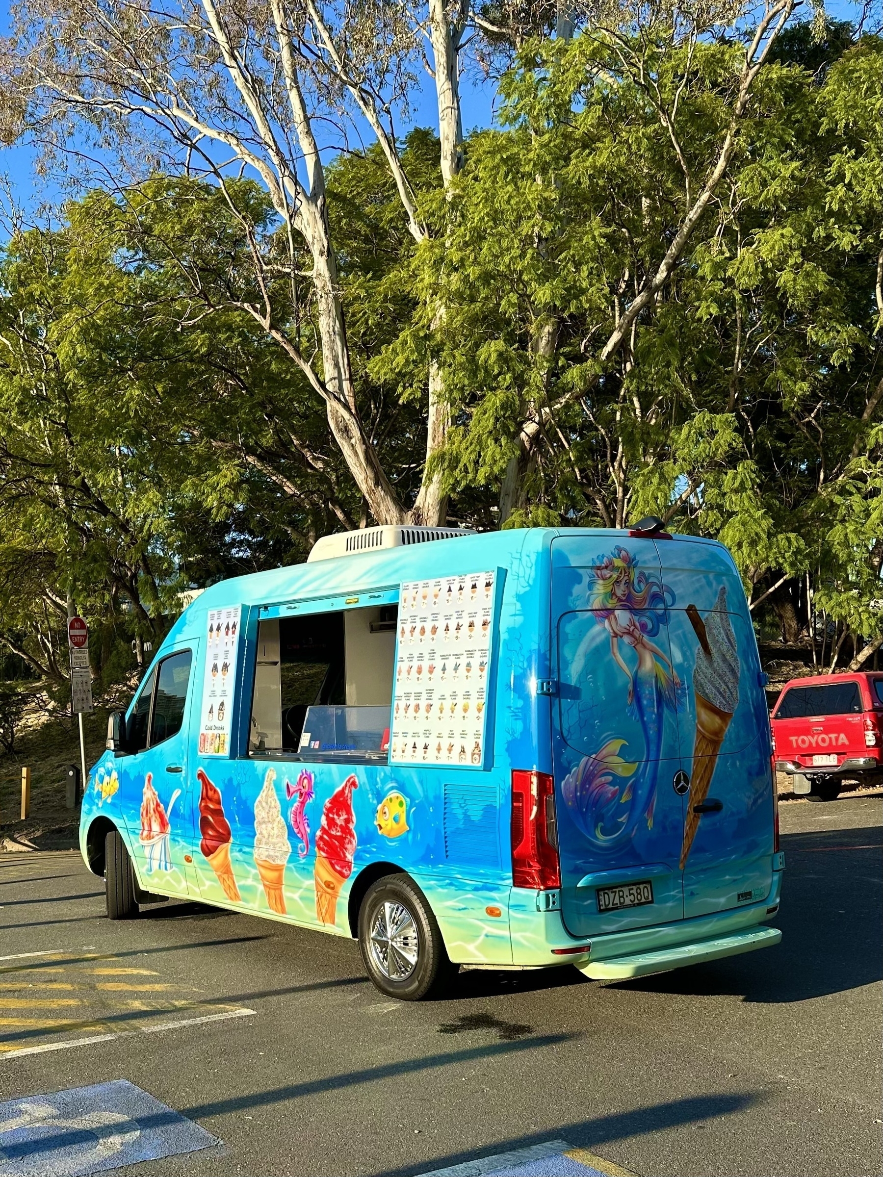 A blue ice cream van parked in front of some trees. The side counter is open and the doors hooked back show the ice creams available. There are ice creams painted in different colours on the truck. The big rear doors have a mermaid and a large ice cream painted on them.