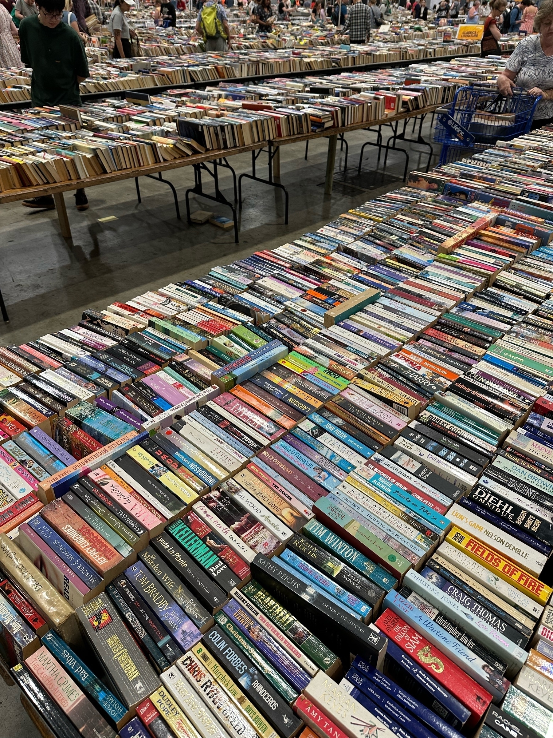 Looking across a hall. Second hand books stacked along trestle tables. Seven rows of paperback books on the table at the front of the photo. Multiple trestle tables stacked similarly in the background.
