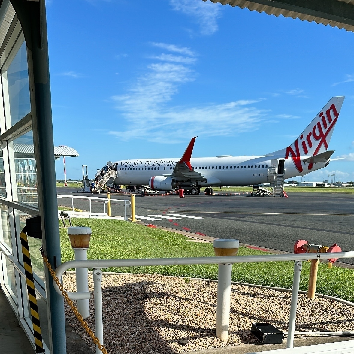 A view of a Virgin Australia plane on the tarmac taken from the boarding area. Entry stairs are at the front and rear of the plane. The plane is white with a red Virgin on the tail. A bright blue sky is visible.