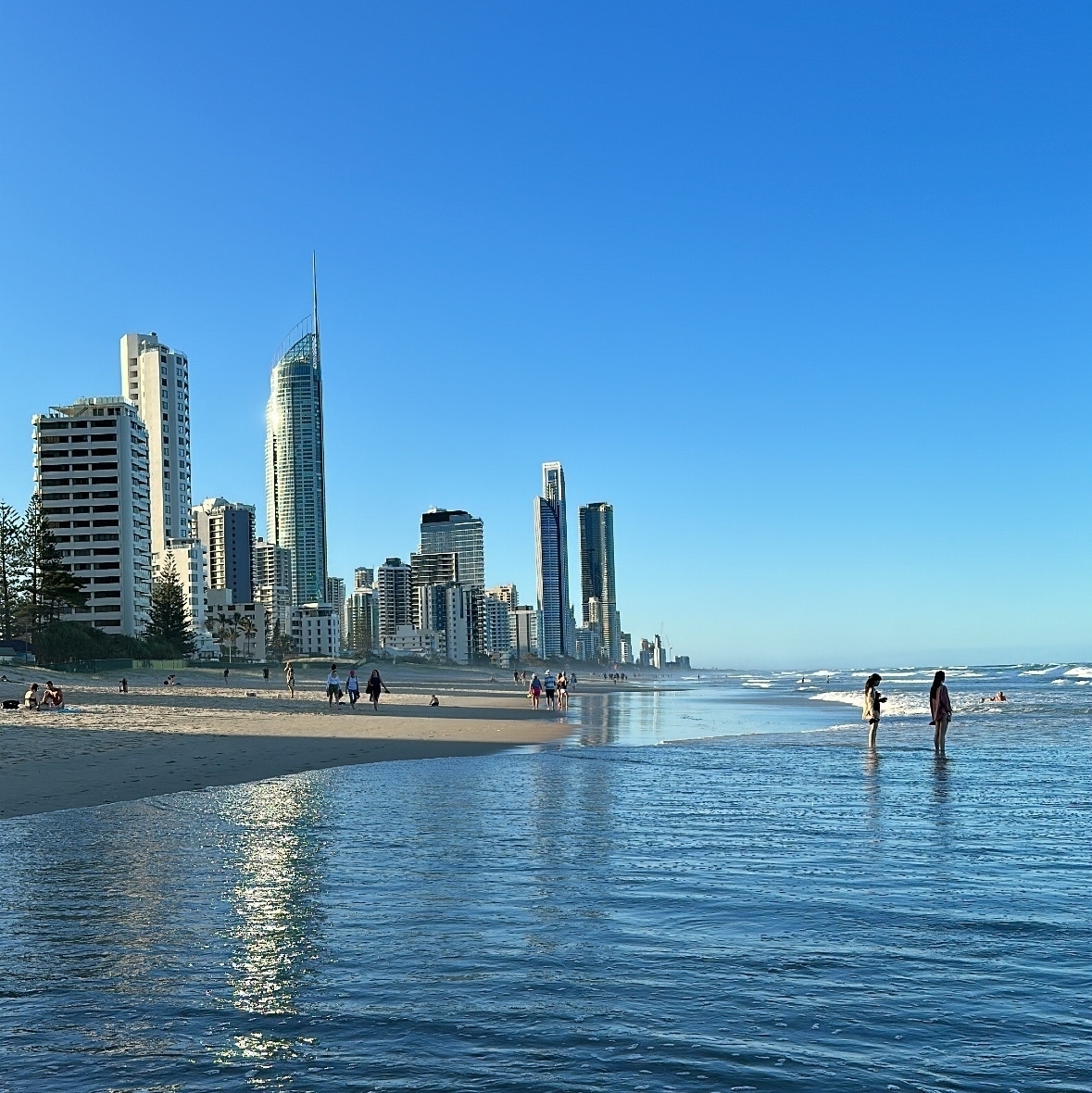 A view along a beach. High rise buildings are to the left of the photo with the beach and water to the right. The photographer is standing in the water that has run up the beach with the surf.