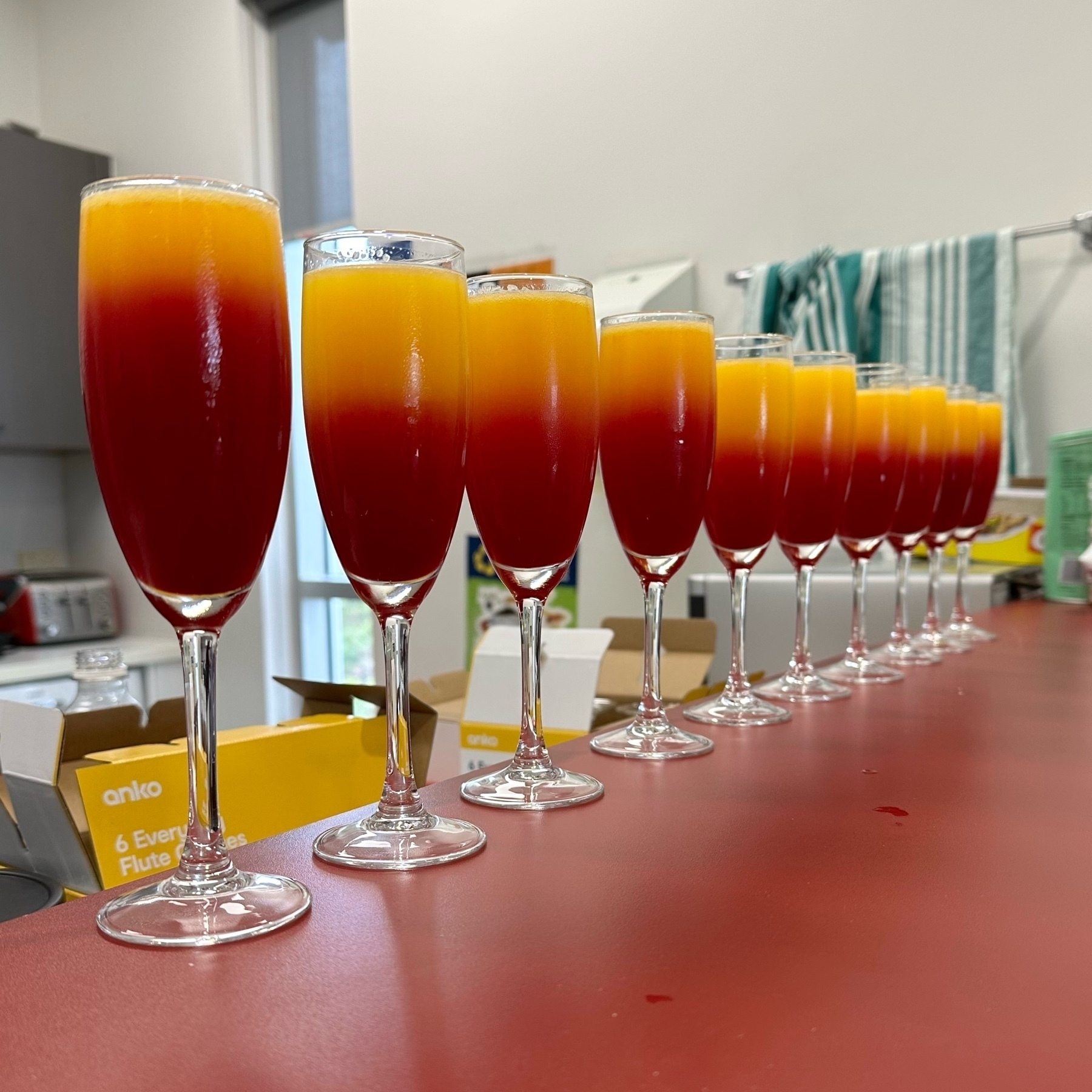 A row of 10 champagne glasses on a red counter top. The drink inside the glasses is coloured red in the bottom half and orange in the top half.