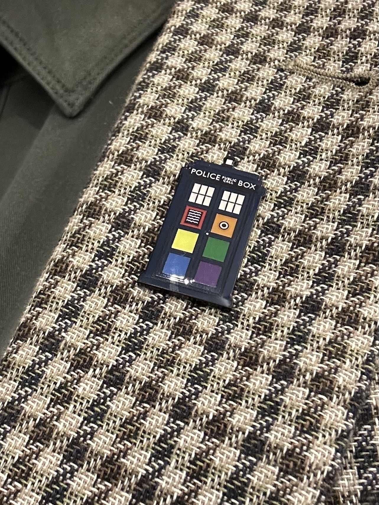 A lapel badge in the shape of the TARDIS police box from the tv show Doctor Who. The badge is blue with rainbow colours in the panels of the Police Box doors. It’s pinned to a green and cream houndstooth pattern blazer.