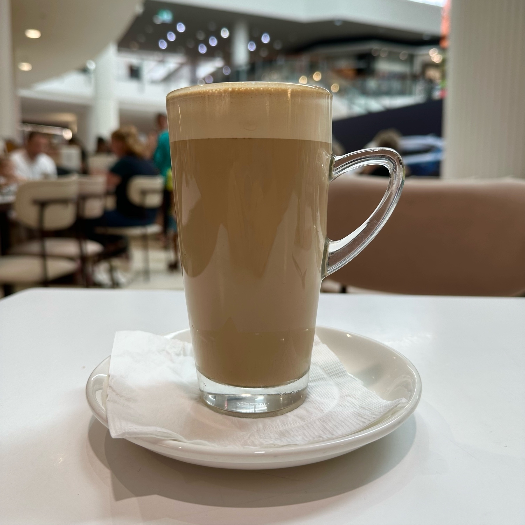 A tall thin glass with a curved glass handle towards the top of the glass. A latte is inside the glass and whiter foam is at the top.