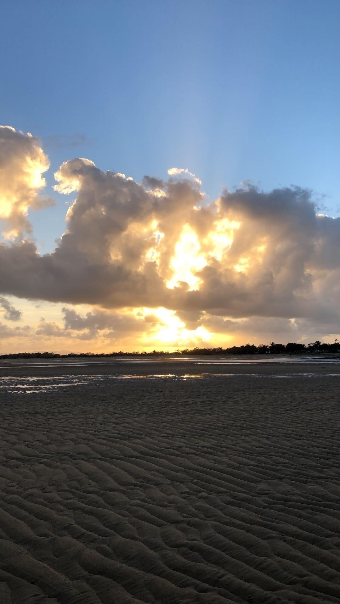A beach view. Sand stretches out in front of the photographer with patches of water where the tide is still running out. The sky has a band of cloud with the sun flaring a yellow orange behind it.