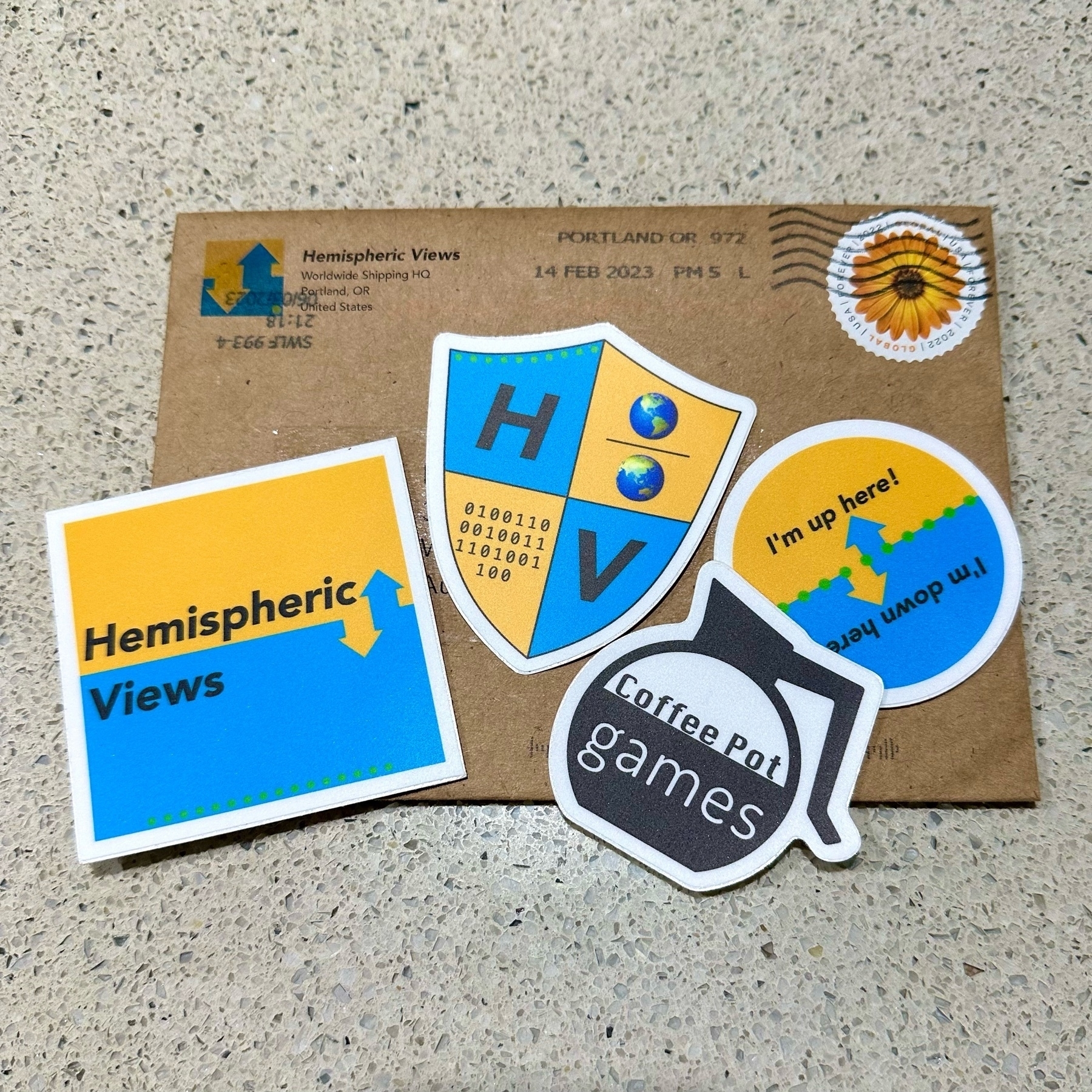 A brown envelope postmarked Portland Oregon on a stone bench top. On top of the envelope are three yellow and blue stickers of different shapes with “Hemispheric Views” on them plus one black and white sticker in the shape of a coffee pot which has “coffee pot games” inside the pot.