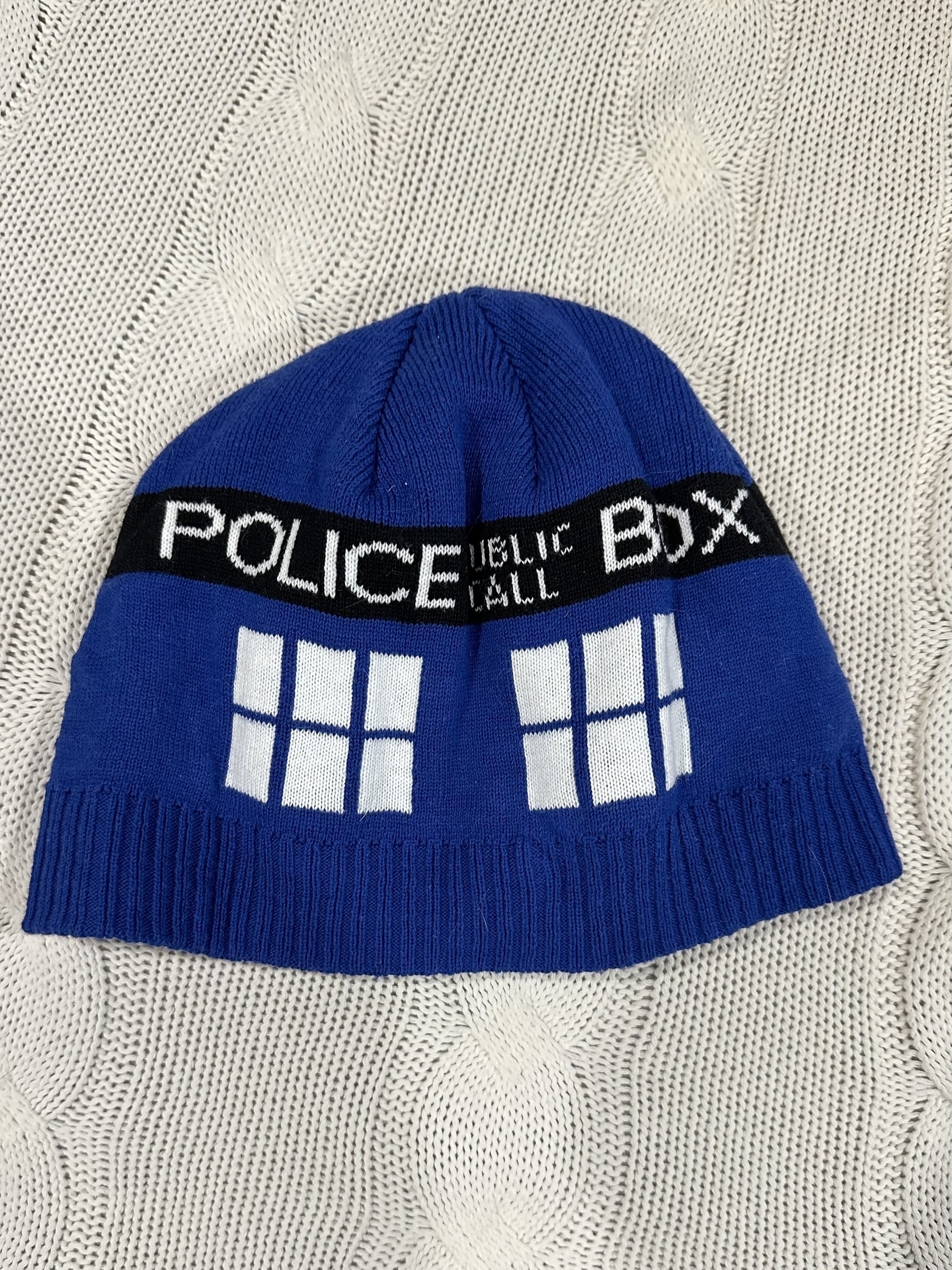 A beanie that looks like the TARDIS from the tv show Doctor Who. It is coloured blue with white windows and a black stripe above them that reads “Police Public Call Box”. 