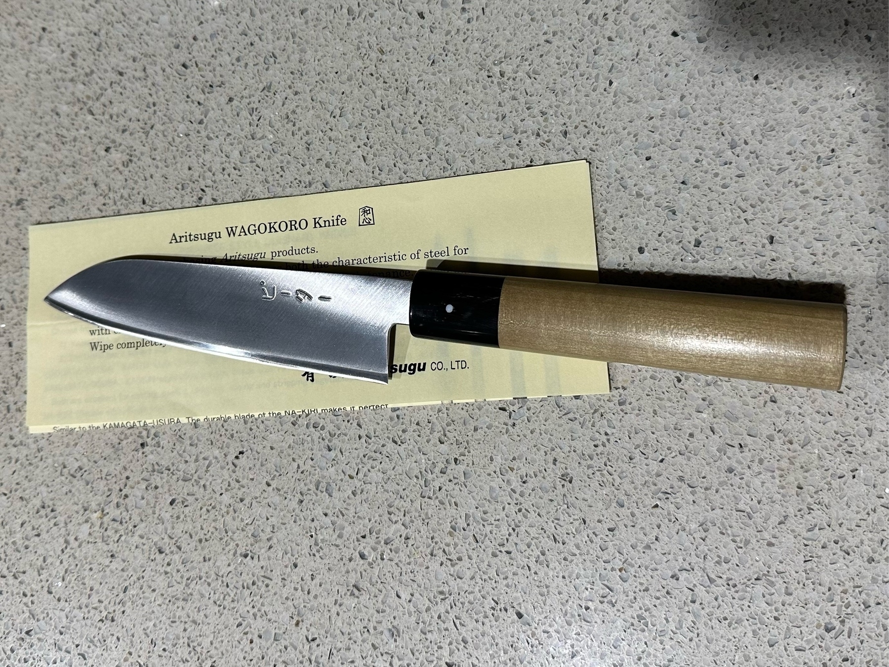 A knife with a wooden handle on a benchtop. The paper under the knife says Aritsugu WAGOKORO knife.
