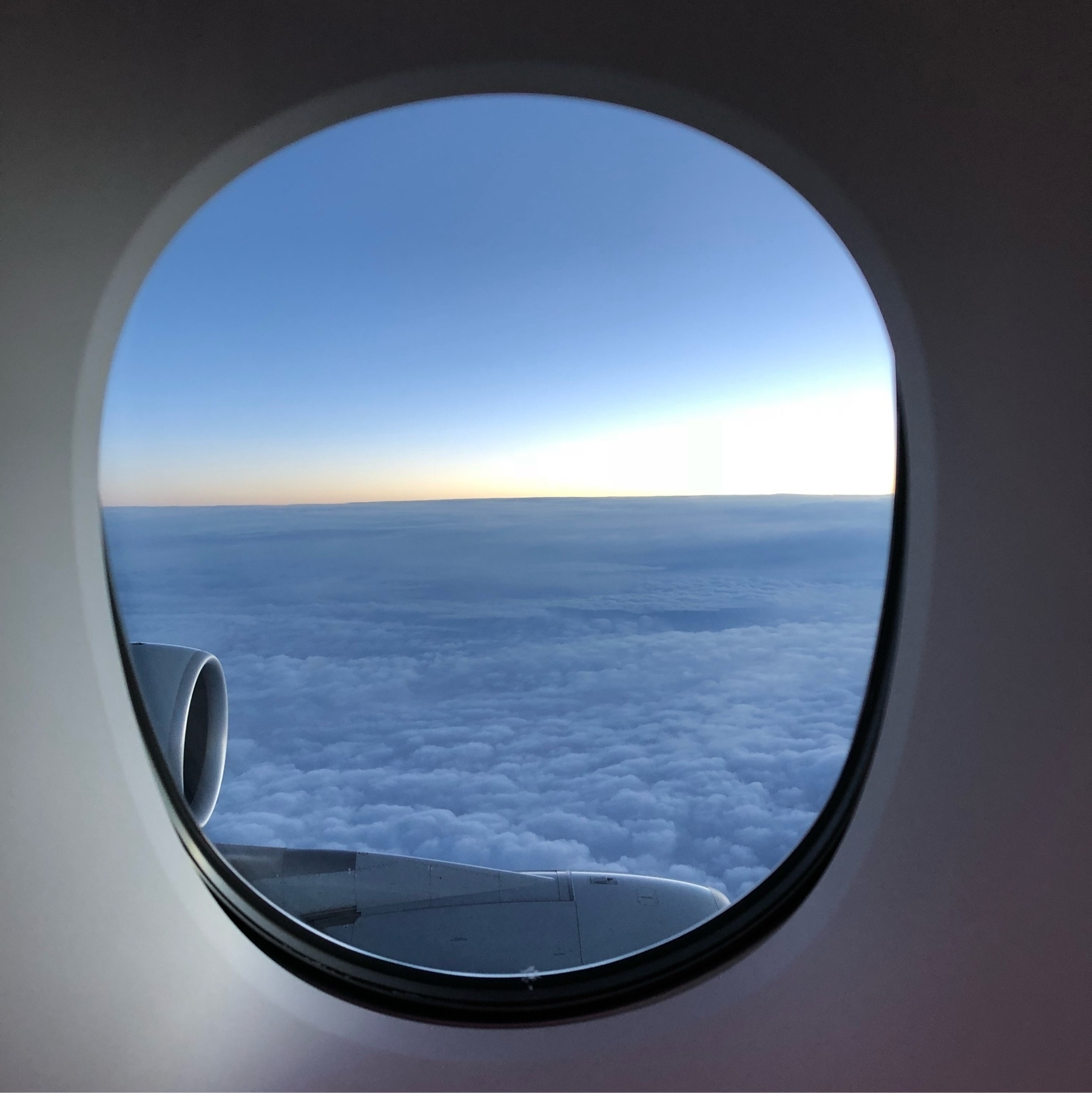 A view looking out of a plane window towards the horizon. A small part of the engine is visible to the left and bottom and the bottom half of the view are white clouds and the upper half blue sky with a bright stripe of sunlight separating the two.