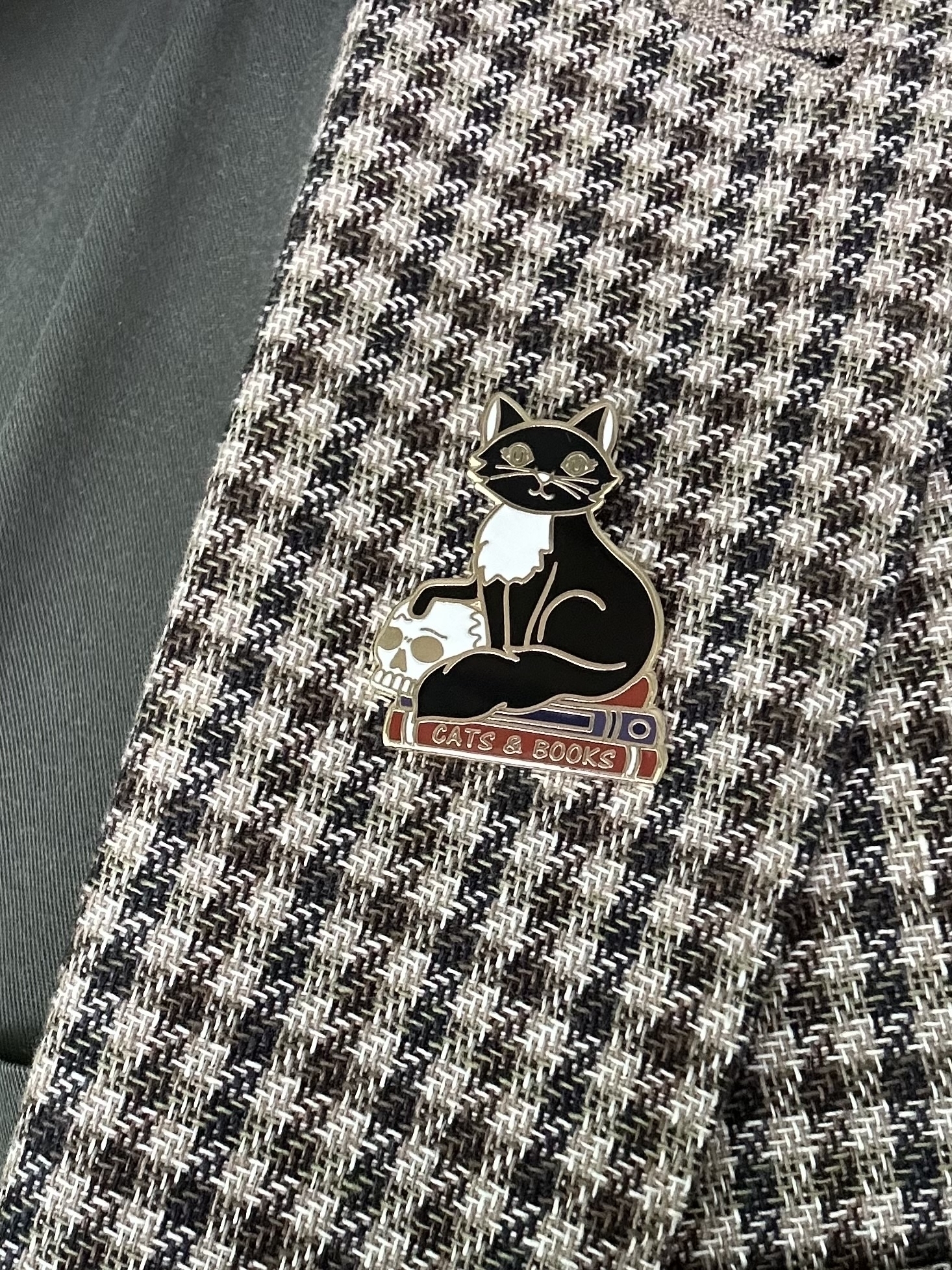 An enamel lapel badge pinned on a green check sports jacket. The badge is a black cat sitting on books with one paw resting on a small white skull. The bottom book is red and has “Cats & Books” in gold lettering on the spine.