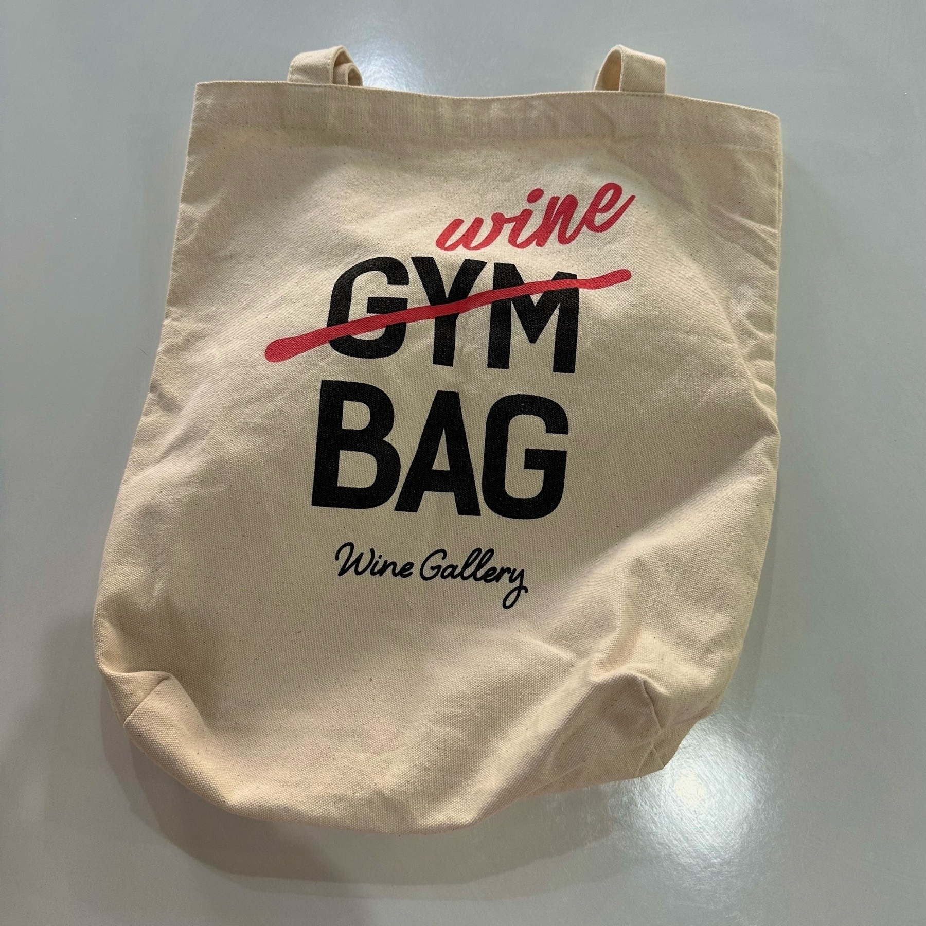 A white cotton bag. Printed on one side is "GYM BAG" with a red line through gym and wine written in red above it. In smaller lettering is "Wine Gallery".