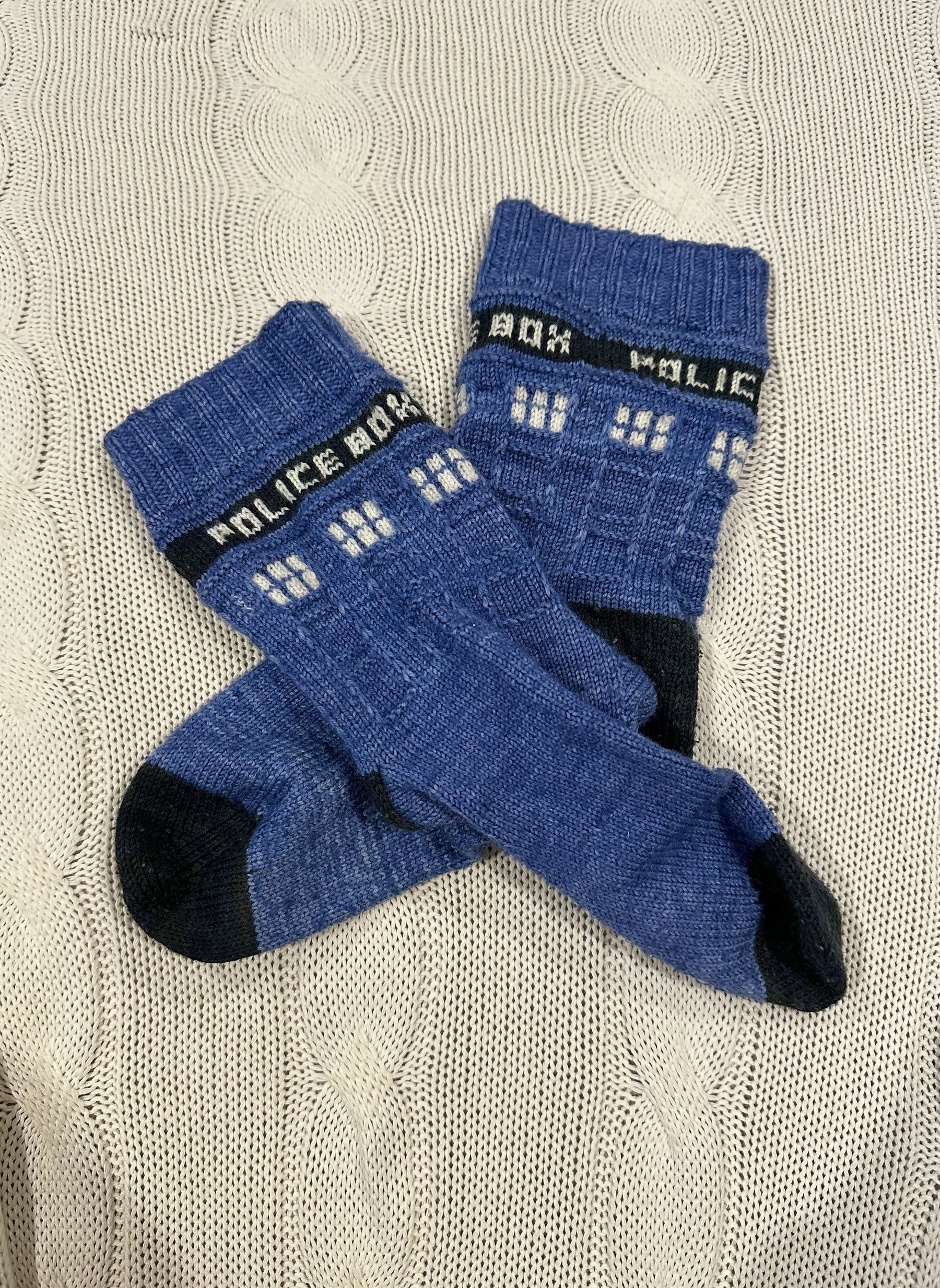 Blue socks knitted to resemble the TARDIS from Doctor Who. “Police Box” is in white lettering inside a black band near the top and white squares underneath are the windows.