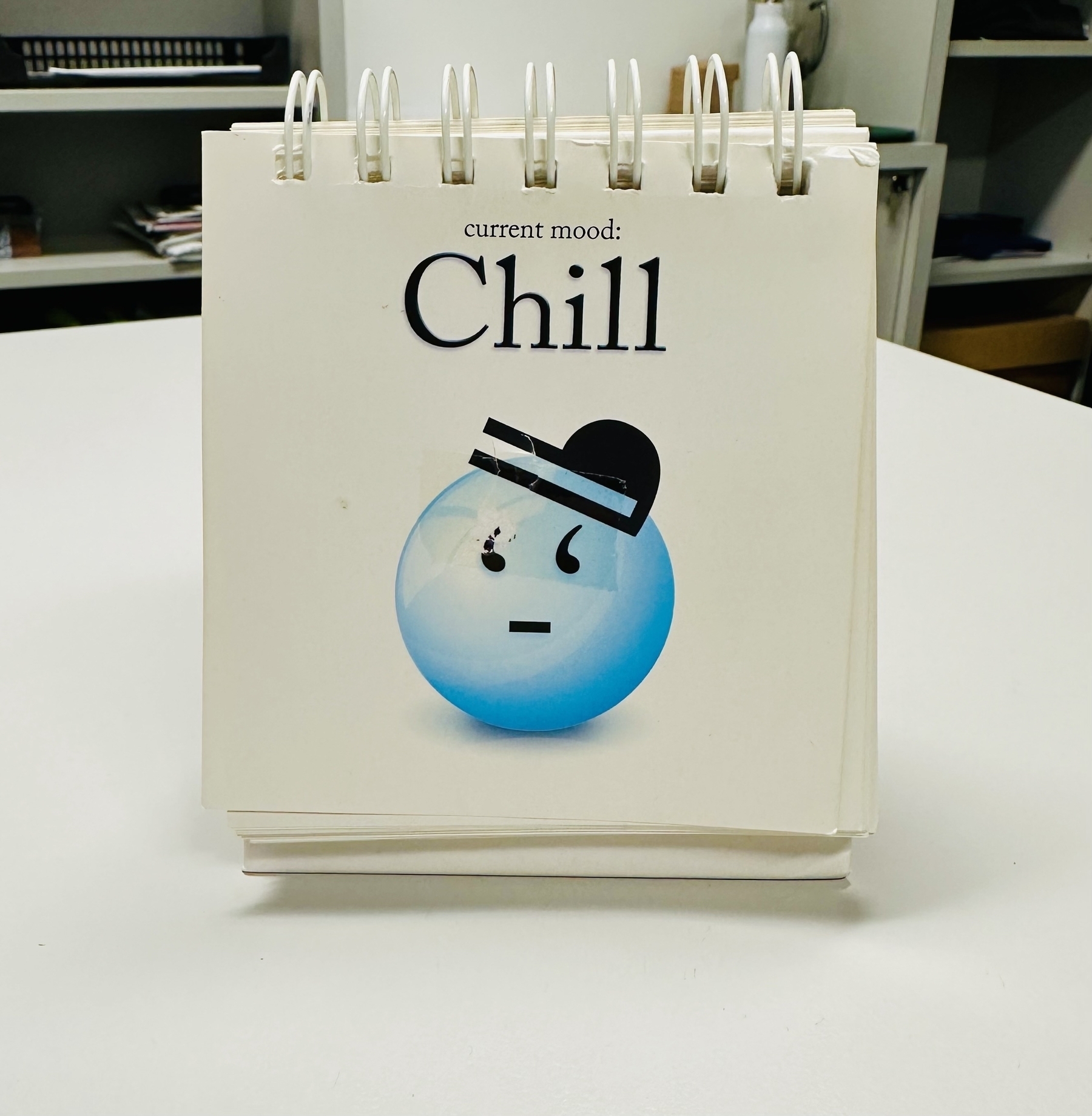 A square sign sitting on a desk. The image says “Current mood:” and then in larger font underneath “Chill”. A blue whimsical emoji face is below that.