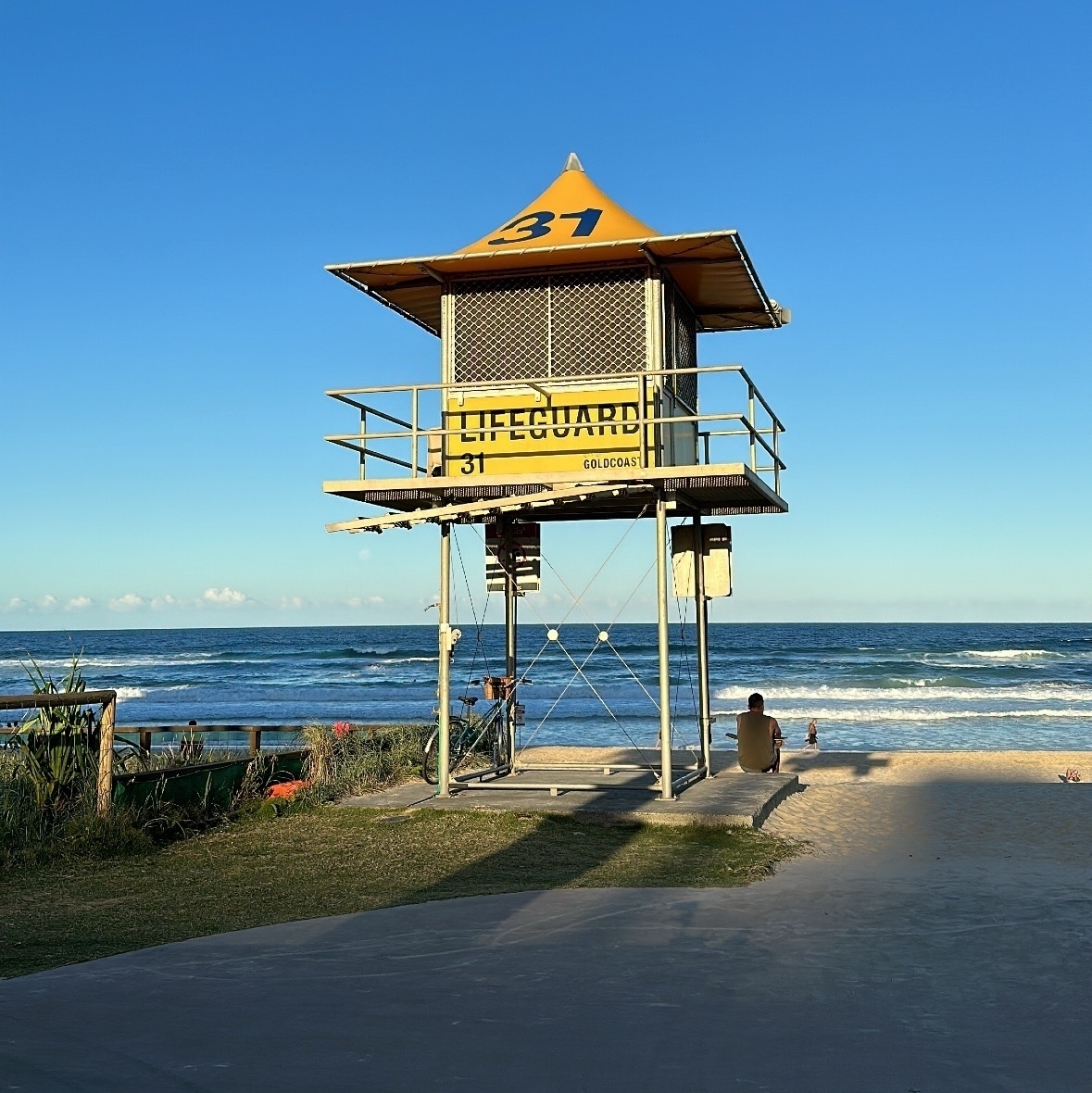 A yellow lifeguard tower with the beach beyond it. A large 31 is painted in blue on the roof of the tower and LIFEGUARD is painted blue on the side. 